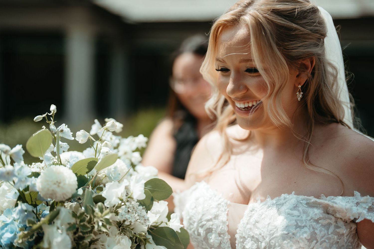 Smiling bride in lace off-shoulder gown holds white and blue floral bouquet with greenery on her wedding day.