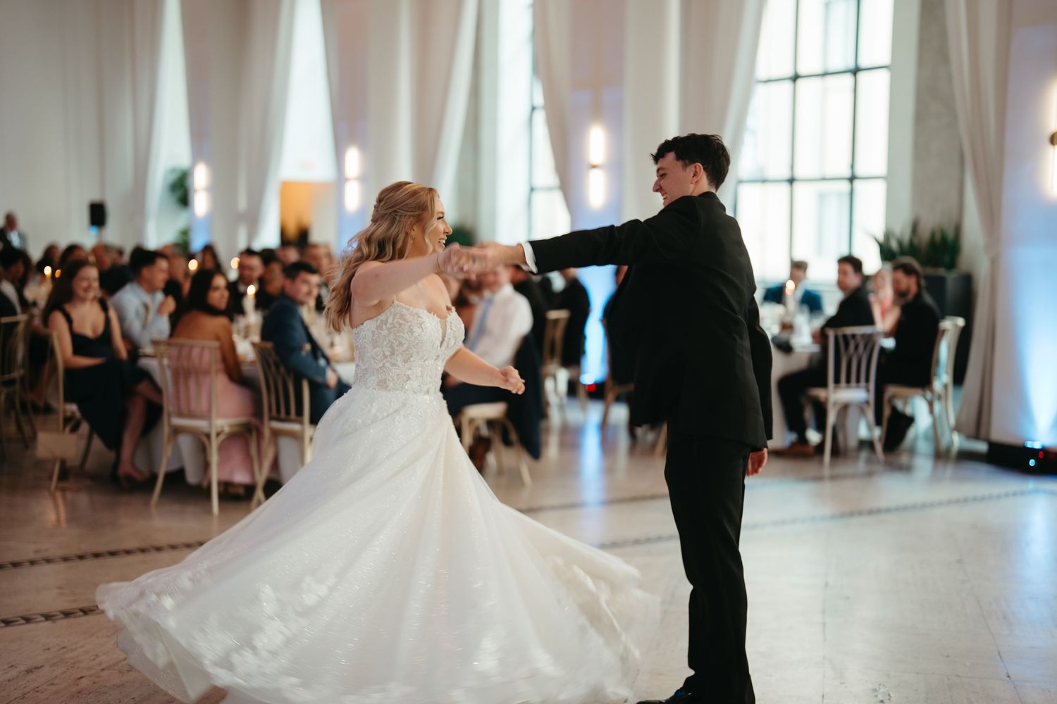 Bride and groom share their first dance in an elegant ballroom with chandelier lighting and guests seated at tables.