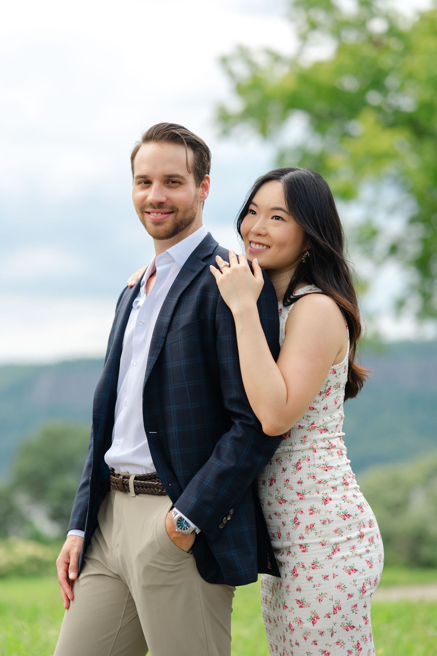 Couple posing outdoors, man in navy blazer and woman in floral dress, smiling with green hills behind them in Untermyer Gardens.