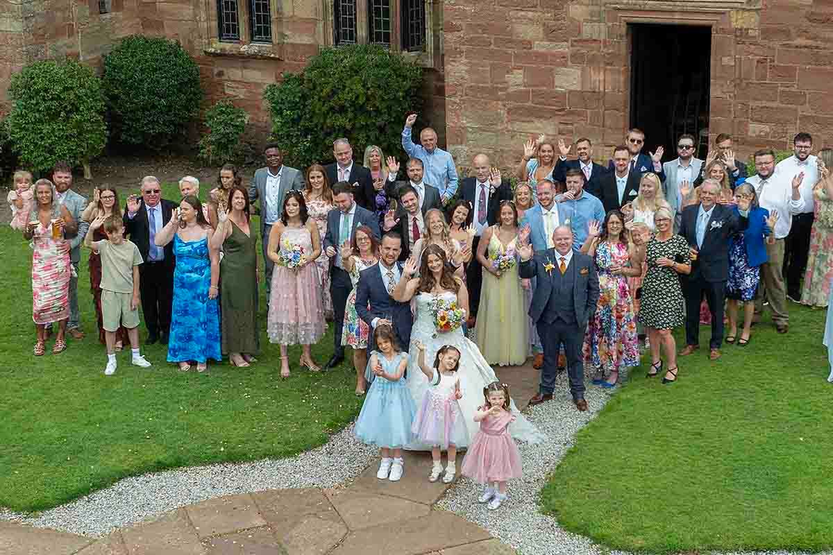Large wedding party group photo outdoors by brick building, bride and groom centered with guests celebrating.