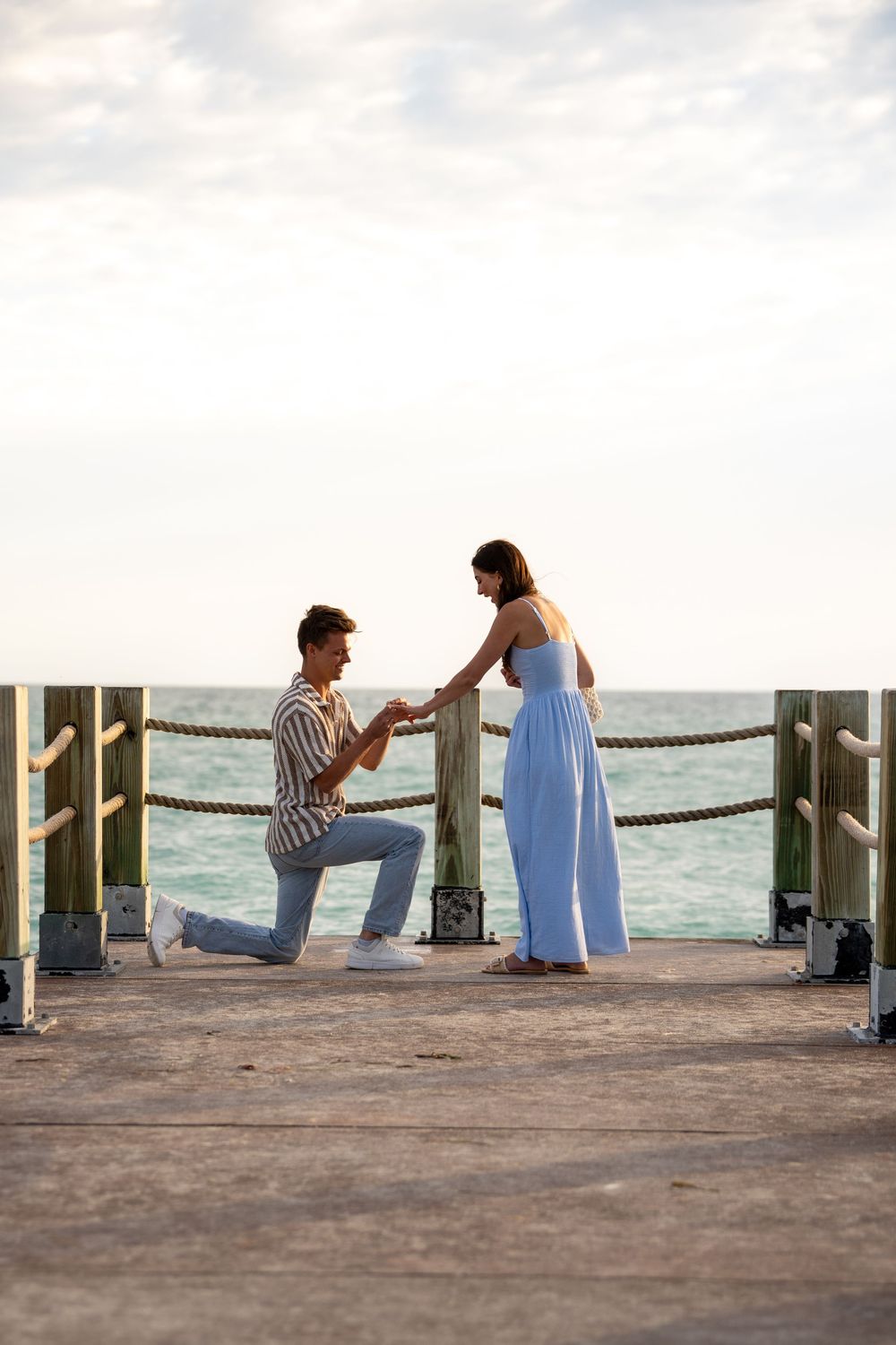 Man proposes on one knee to woman in blue dress on ocean pier at sunset.