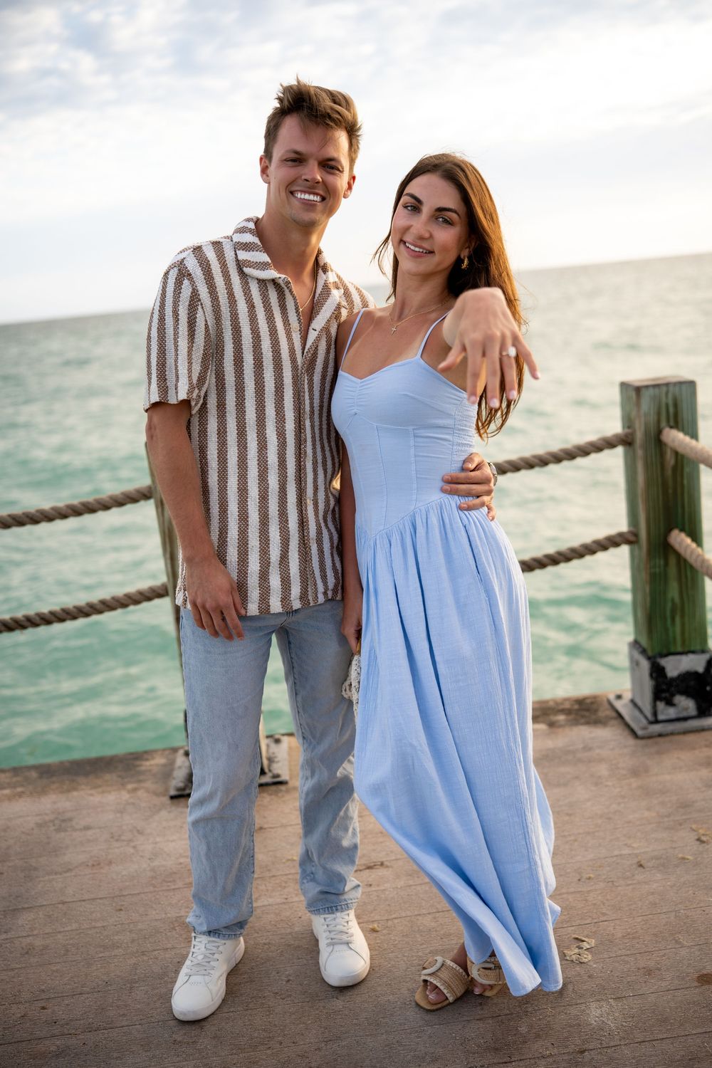 A smiling couple poses on a seaside pier, man in striped shirt and jeans, woman in light blue maxi dress.