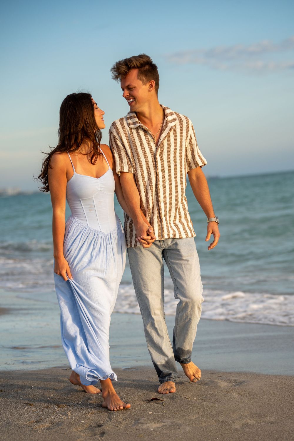 Happy couple holding hands and walking barefoot on a sandy beach at sunset, woman in blue dress, man in striped shirt.