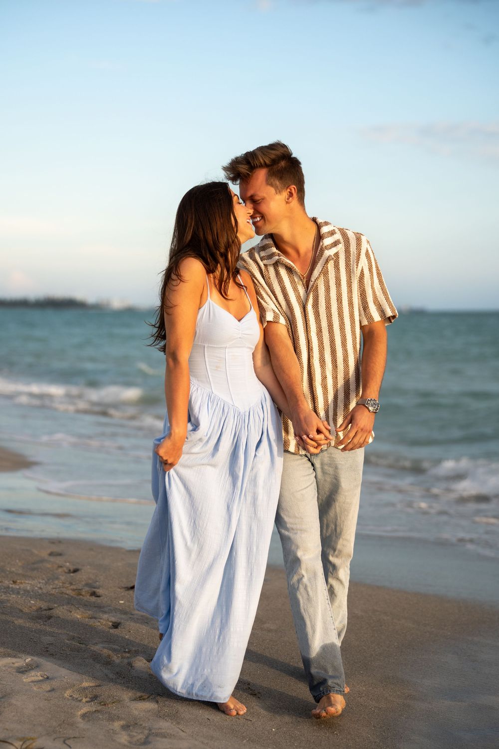 A romantic couple walks hand-in-hand along a sandy beach at sunset, smiling at each other.