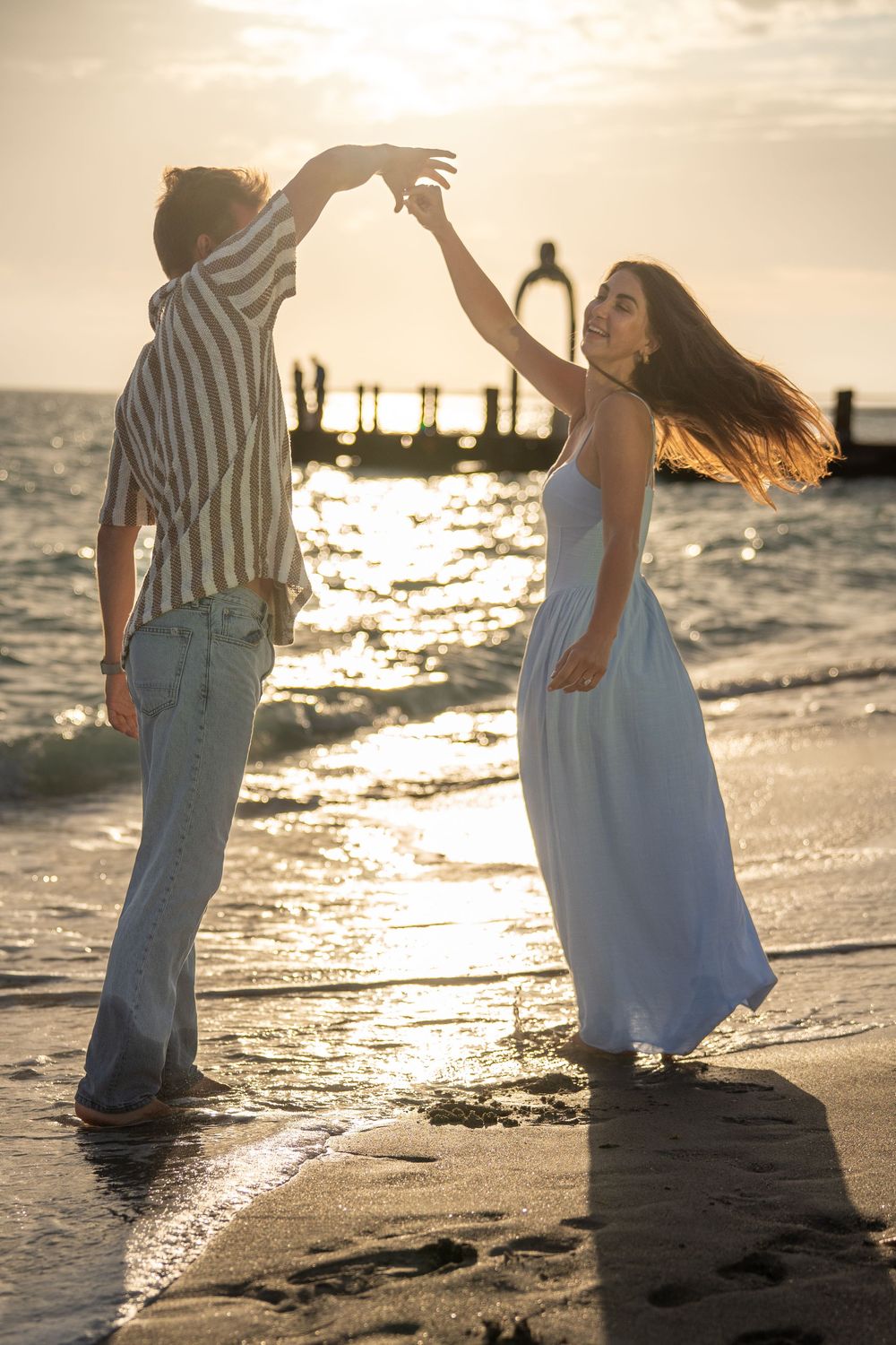 Couple dancing romantically on a beach at golden sunset, woman in blue dress twirling near ocean waves.