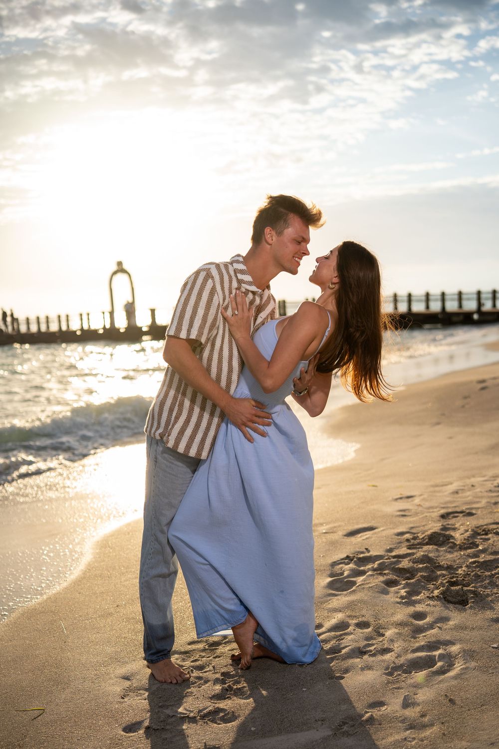 Couple embracing on a sandy beach at golden sunset, woman in blue dress, waves and pier in background.