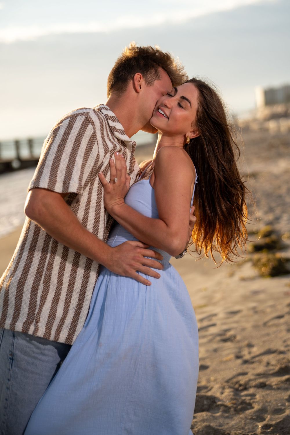 Romantic couple embracing on a sandy beach at sunset, woman in blue dress smiling as man kisses her cheek.