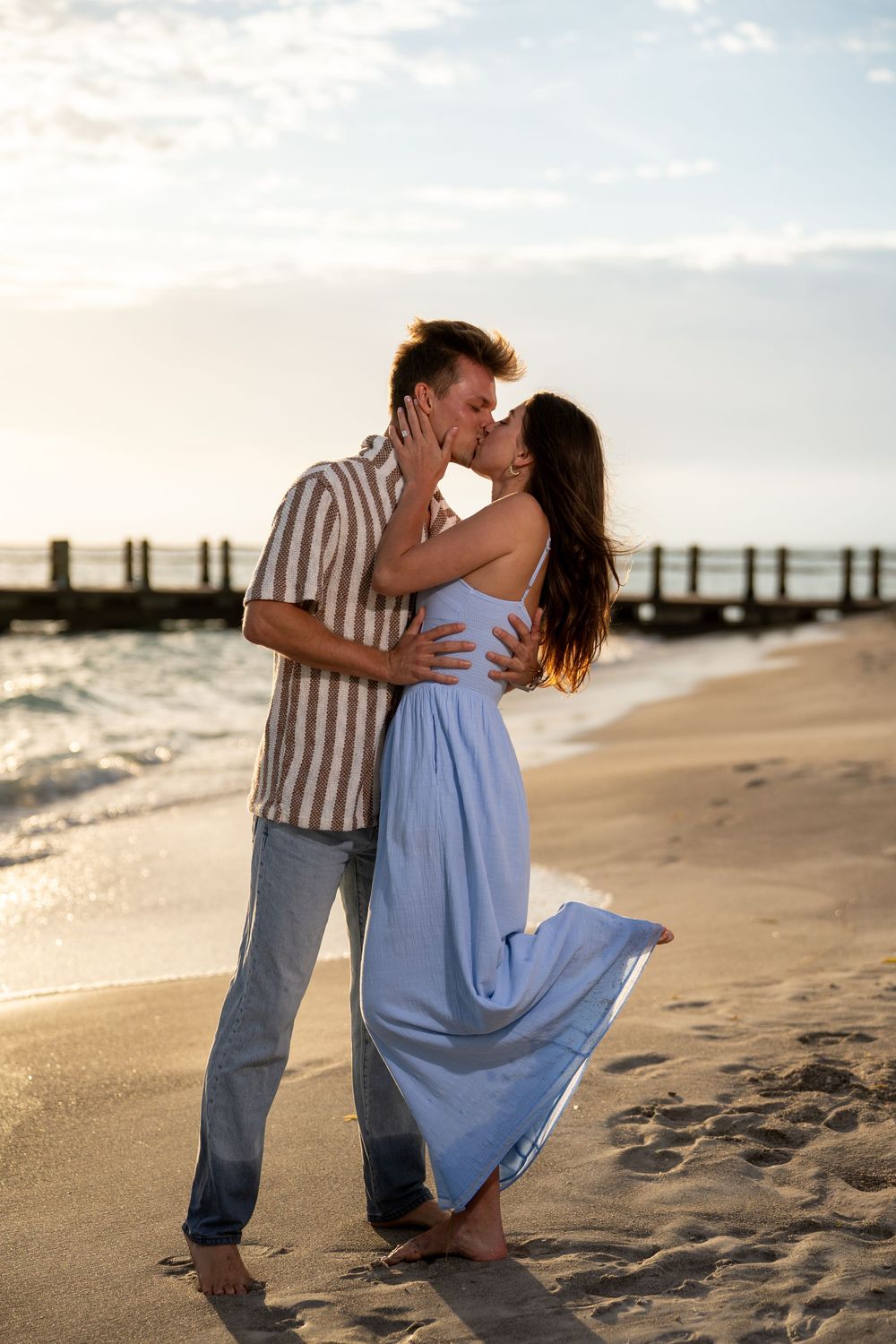 Couple kissing on a sandy beach at sunset, woman in flowing blue dress, man in striped shirt and jeans.
