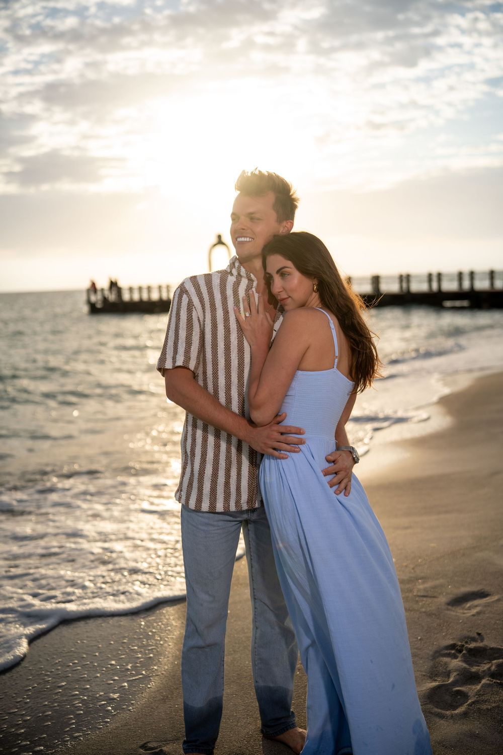 Couple embracing on a sandy beach at golden sunset, woman in blue maxi dress, man in striped shirt, ocean pier behind.