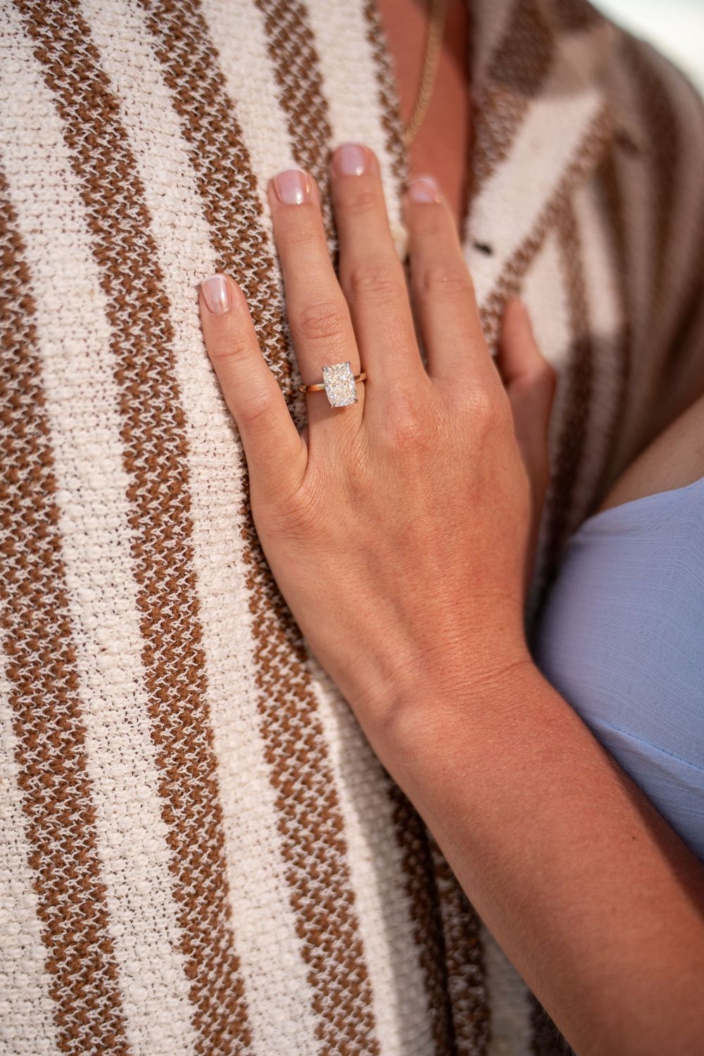 Close-up of a woman's hand wearing a diamond halo engagement ring on rose gold band, resting on a striped knit fabric.