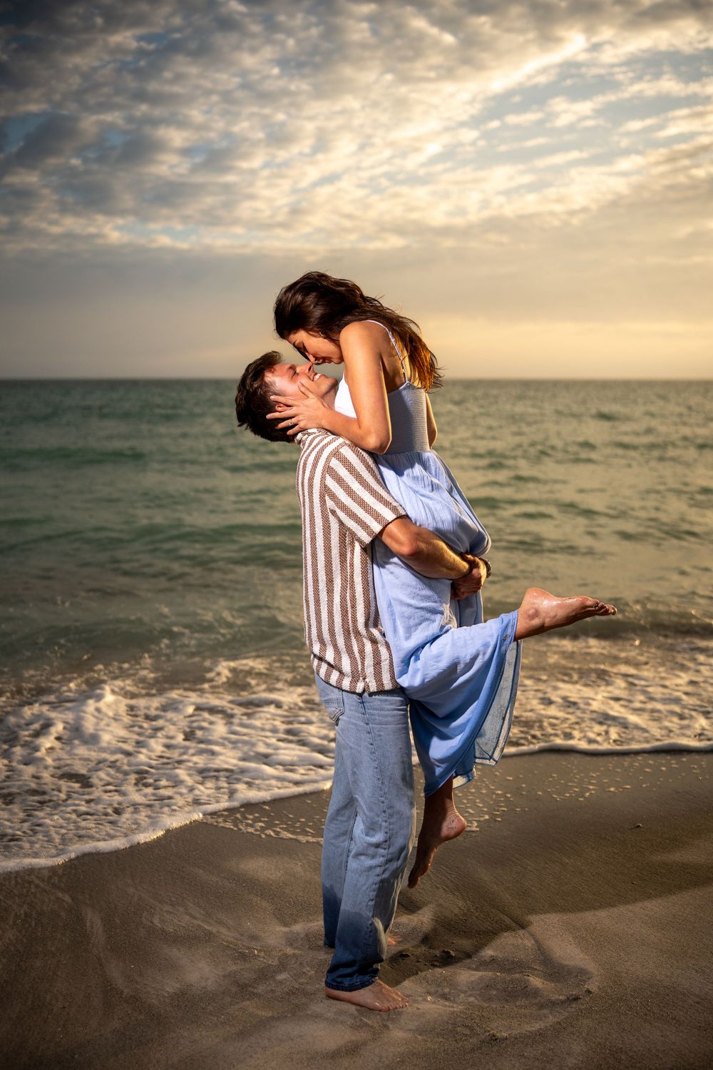 Romantic couple kissing on a sandy beach at sunset, man lifting woman in blue dress near ocean waves.