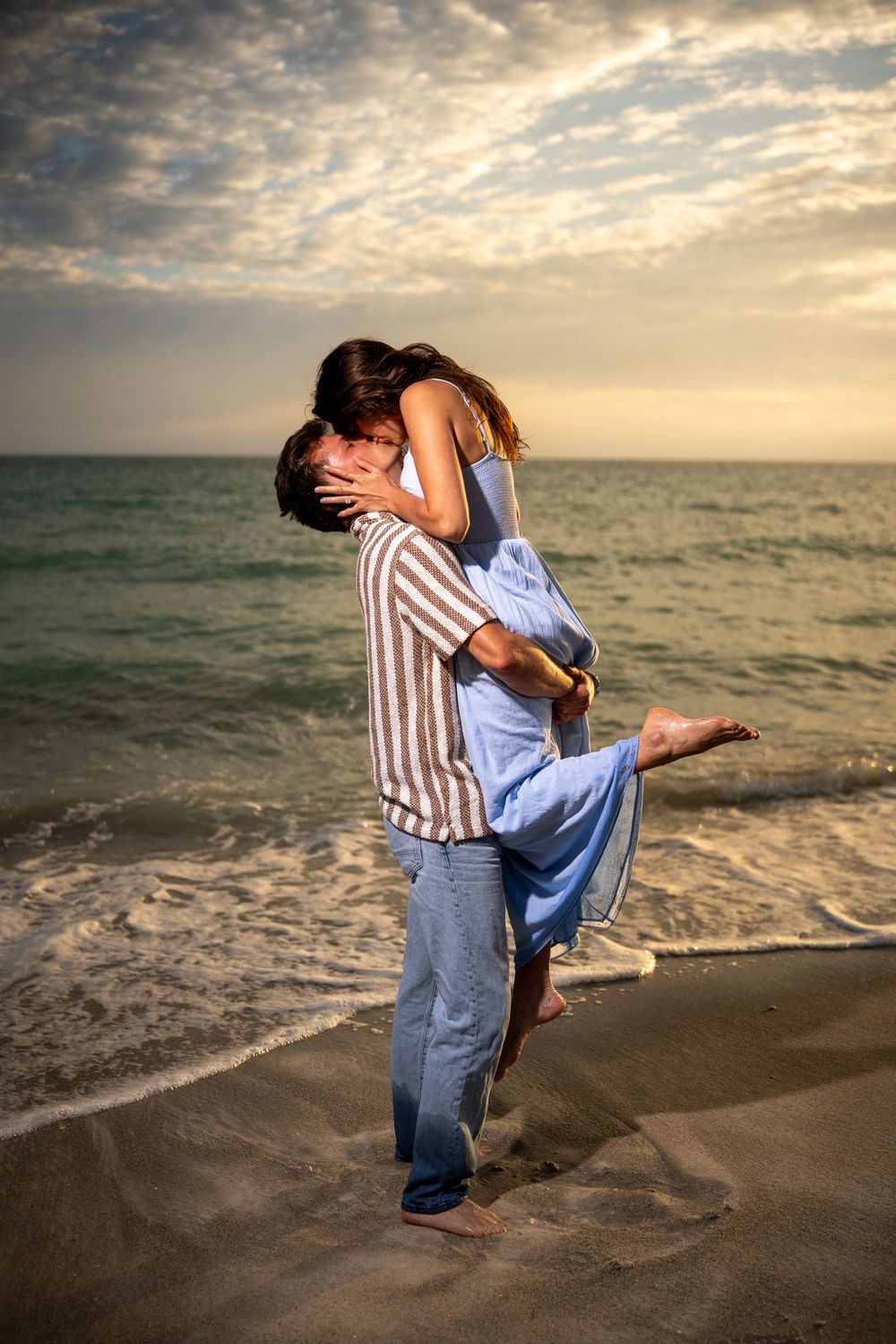 Romantic couple embracing and kissing on a sandy beach at sunset with ocean waves in the background.