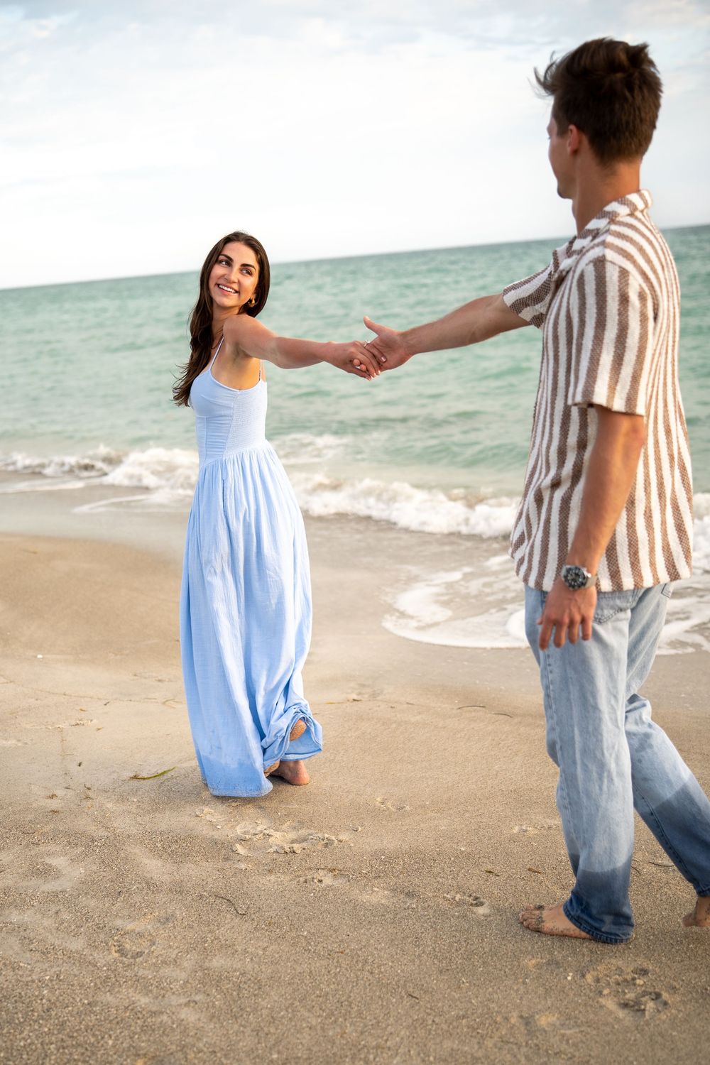 Happy couple holding hands on a sandy beach, woman in blue maxi dress smiling at man in striped shirt.