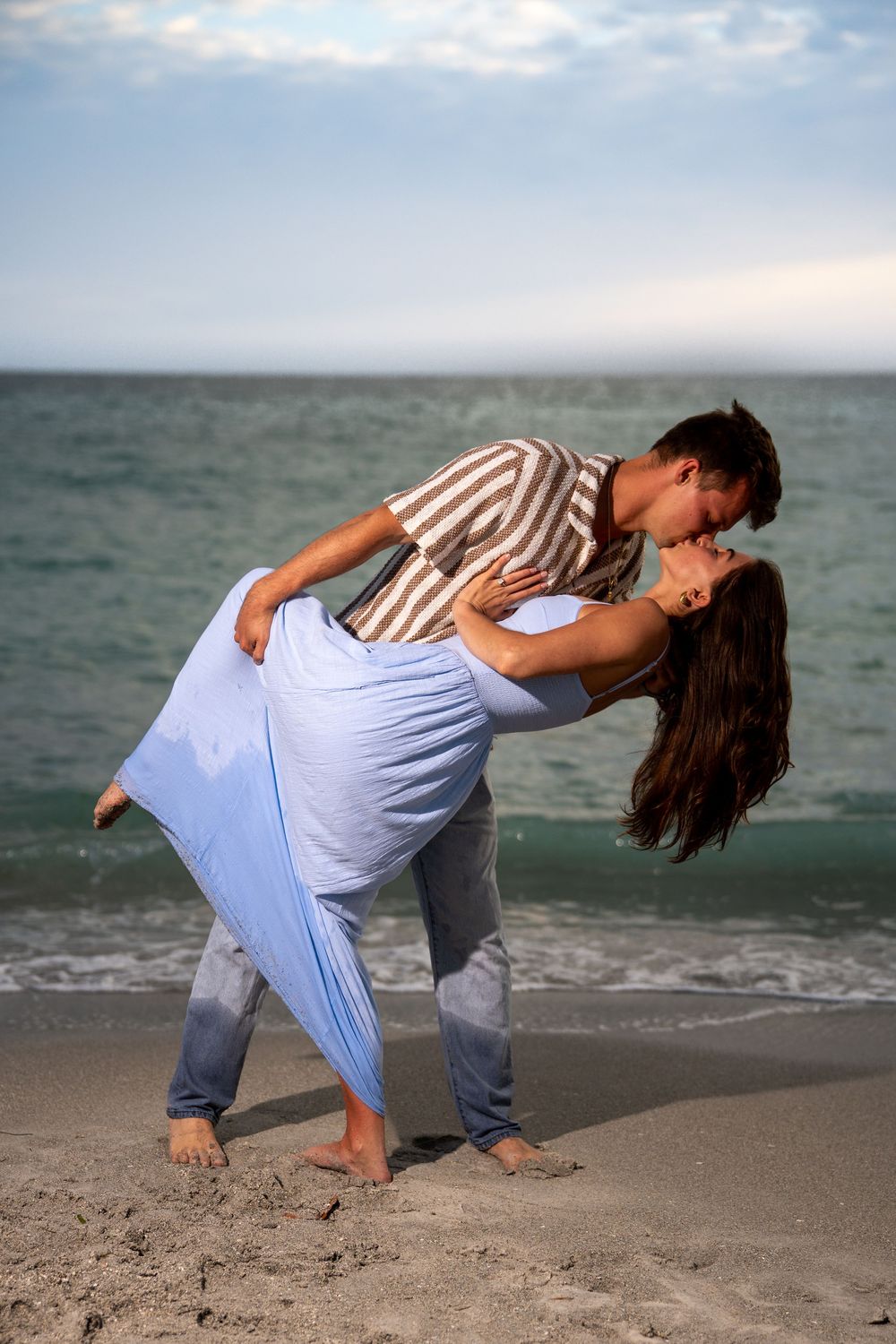 Romantic couple sharing a passionate dip kiss on a sandy beach with ocean waves in the background.