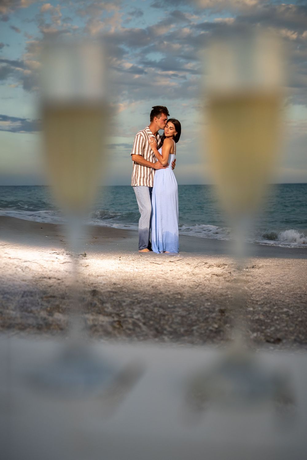 Romantic couple embracing on a beach at sunset with champagne glasses in the foreground.