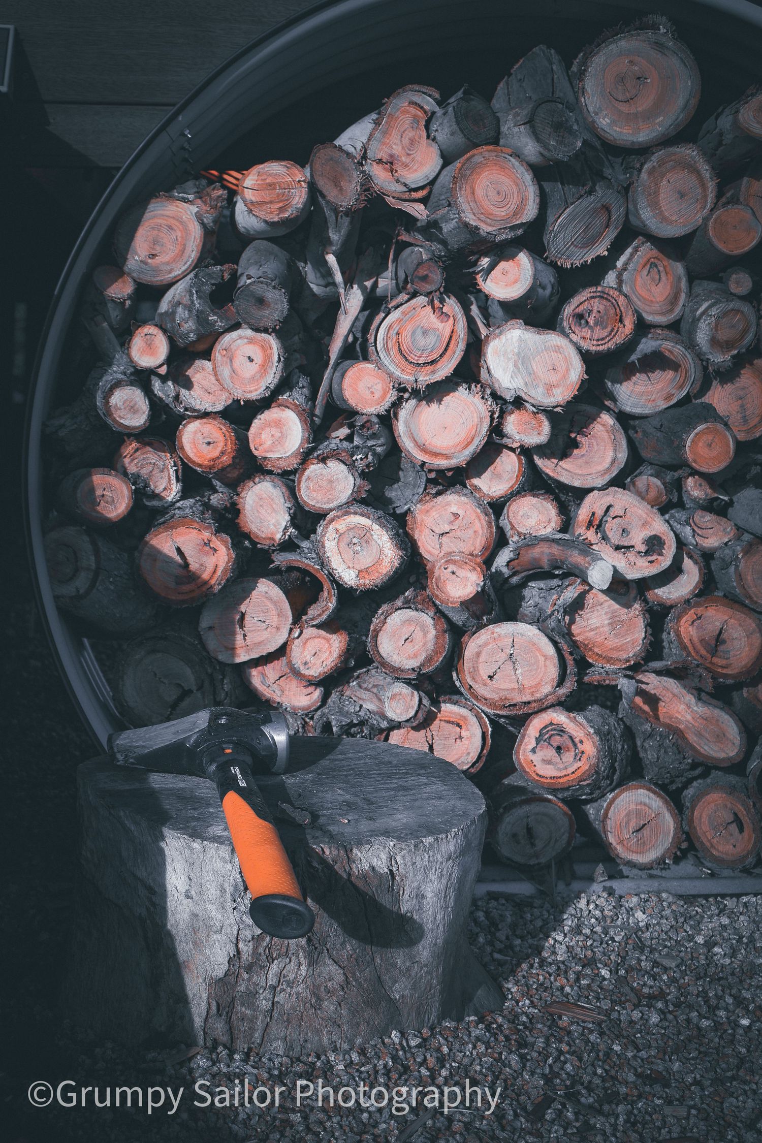 Stacked cut firewood logs with visible rings fill a large round container, with an axe resting on a stump in foreground.