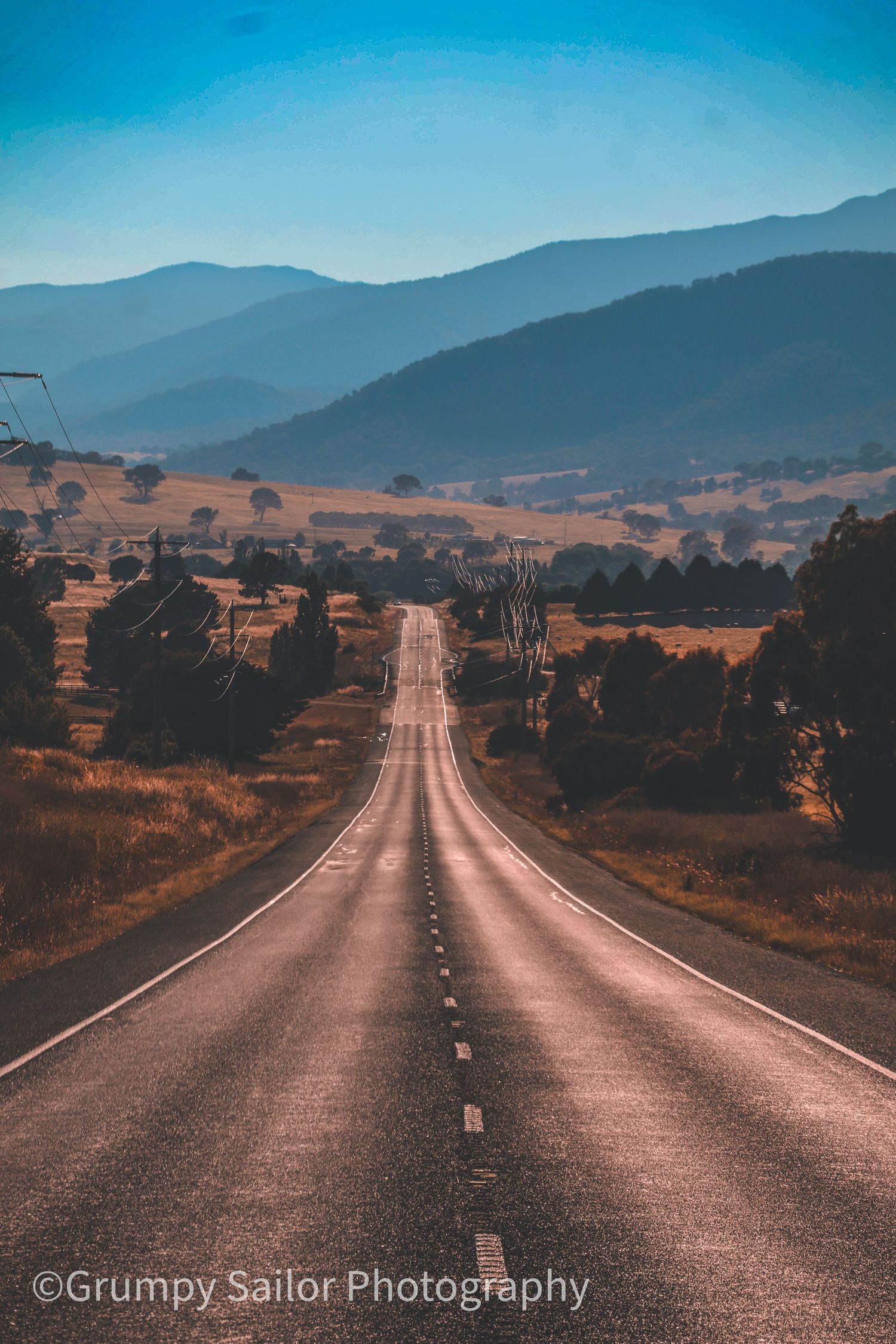 Empty two-lane highway stretching toward misty blue mountains through golden Australian countryside on a clear day.