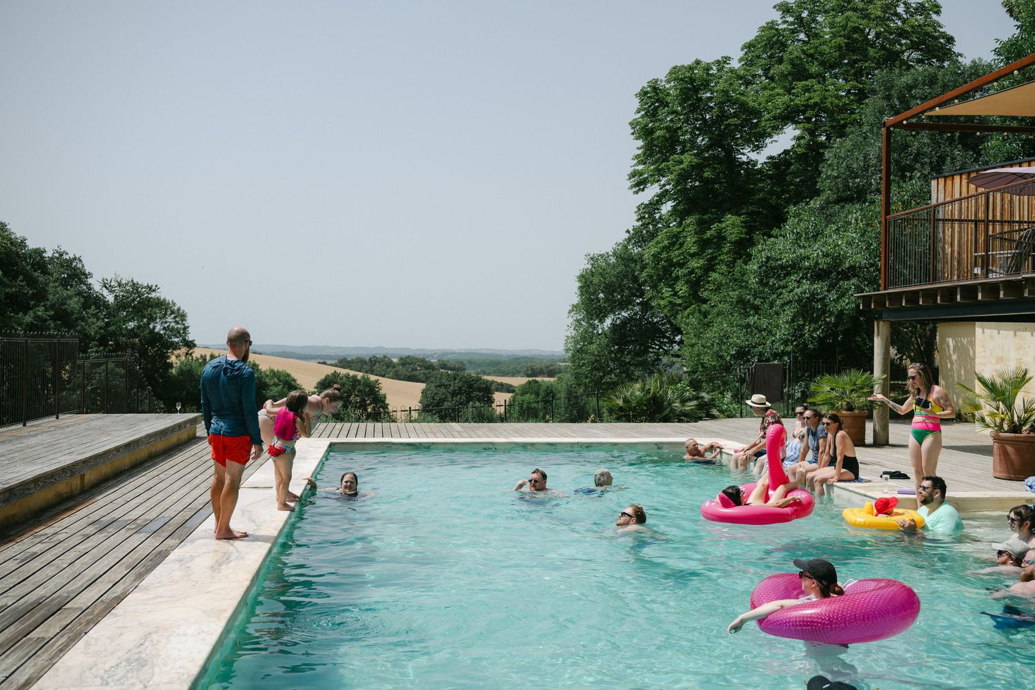 People enjoying a sunny day swimming in an outdoor infinity pool with colorful floats, surrounded by lush countryside views.