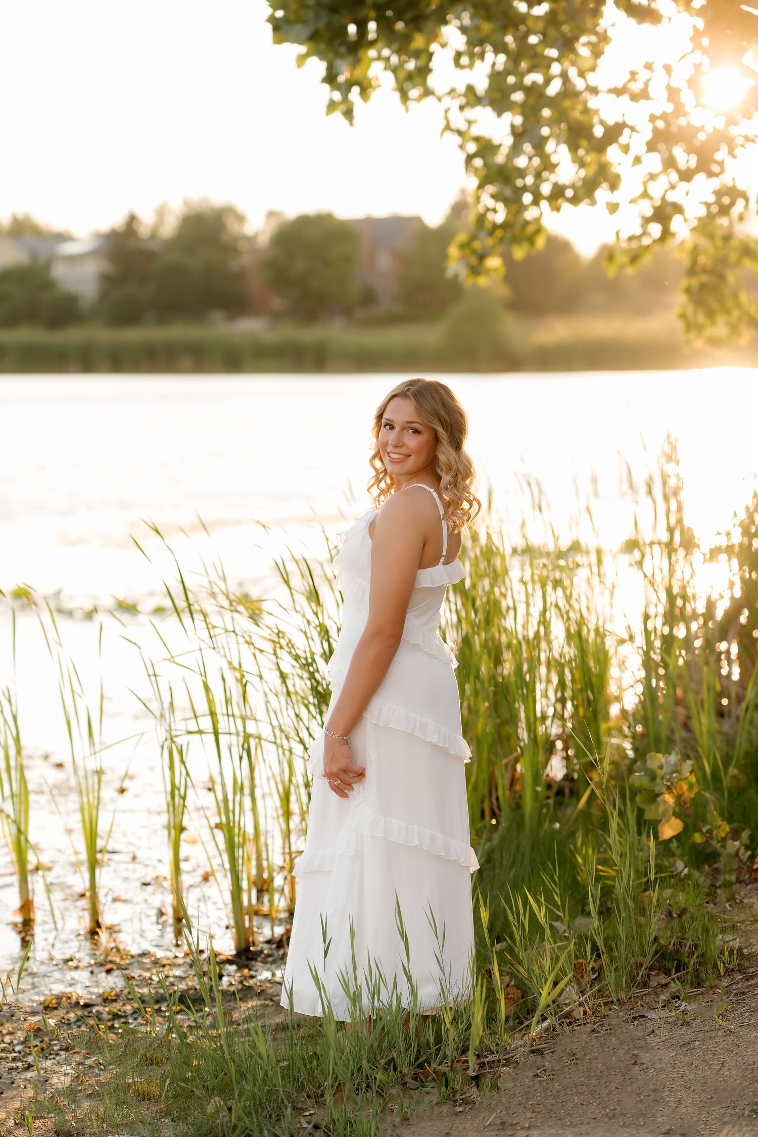 Young woman in white dress standing by a lake at golden sunset, smiling over her shoulder near tall reeds.