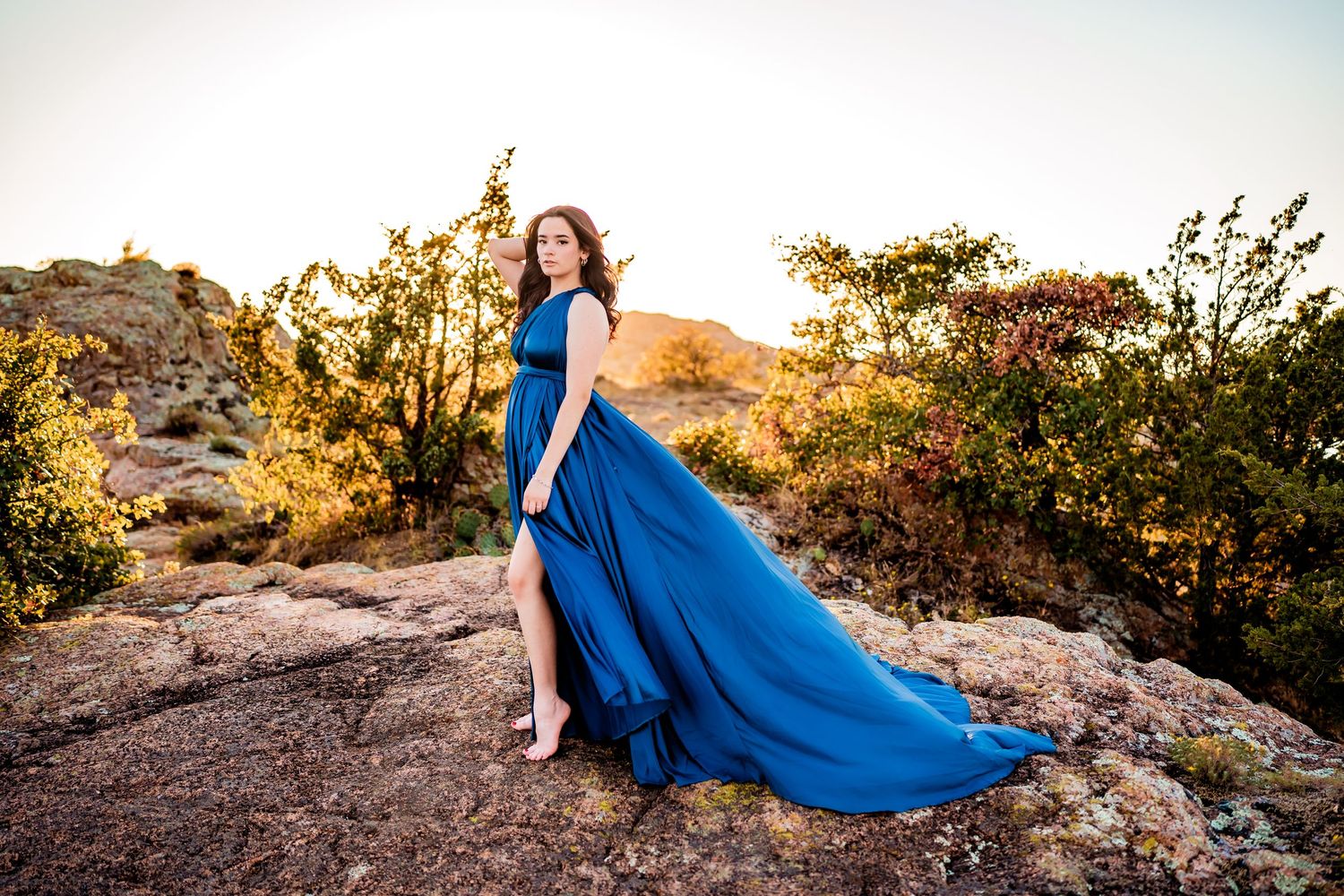 Woman in flowing blue gown poses on rocky desert landscape at golden hour sunset.