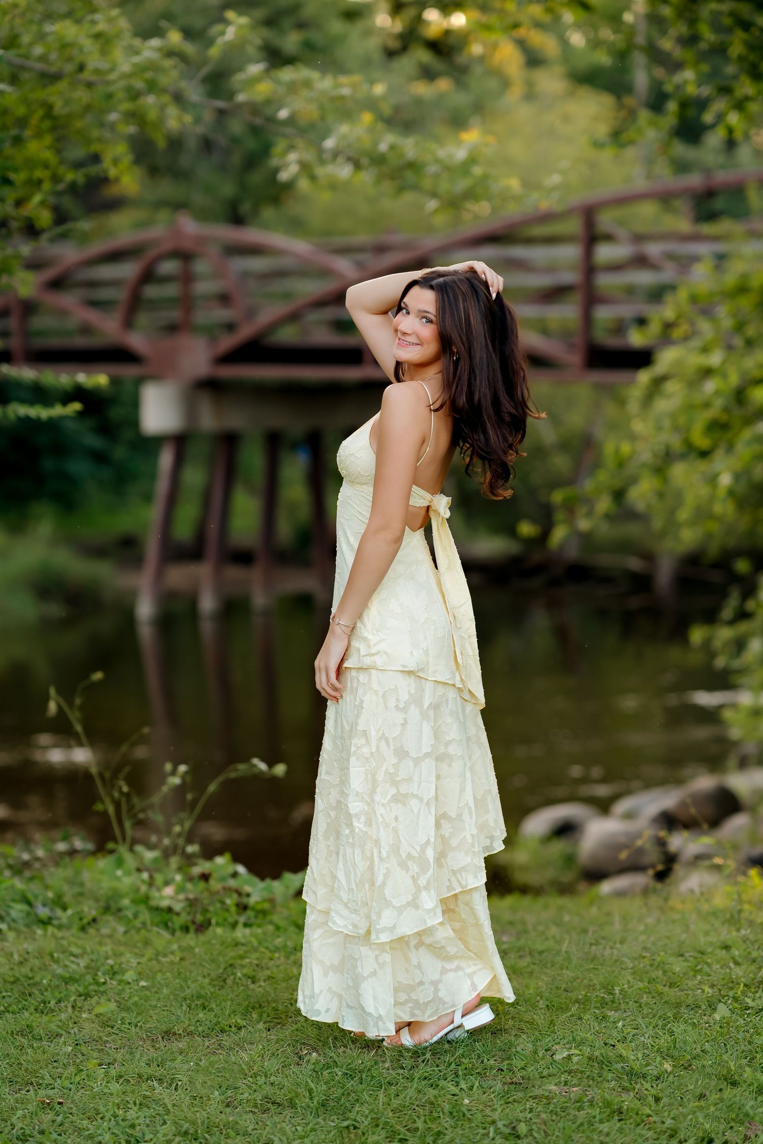 Woman in elegant yellow strapless gown posing outdoors near a rustic bridge and pond surrounded by lush greenery.
