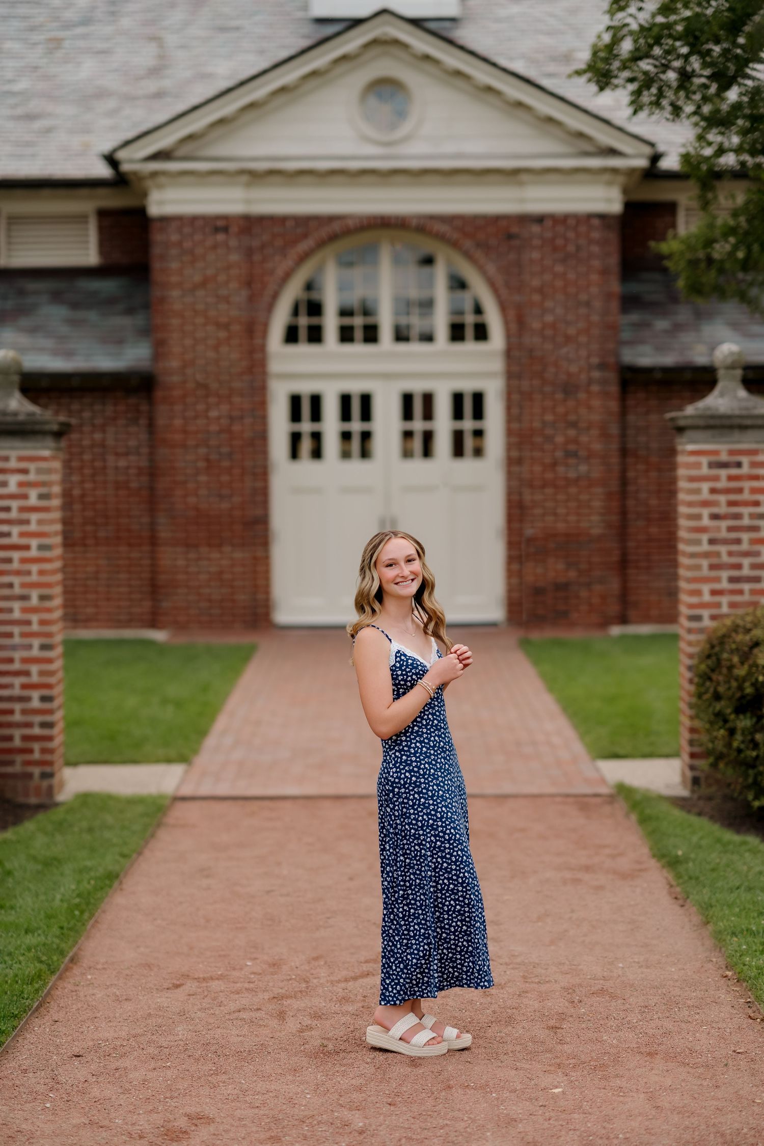 Young woman in navy floral maxi dress smiling on brick pathway in front of elegant red brick building with arched white doors.
