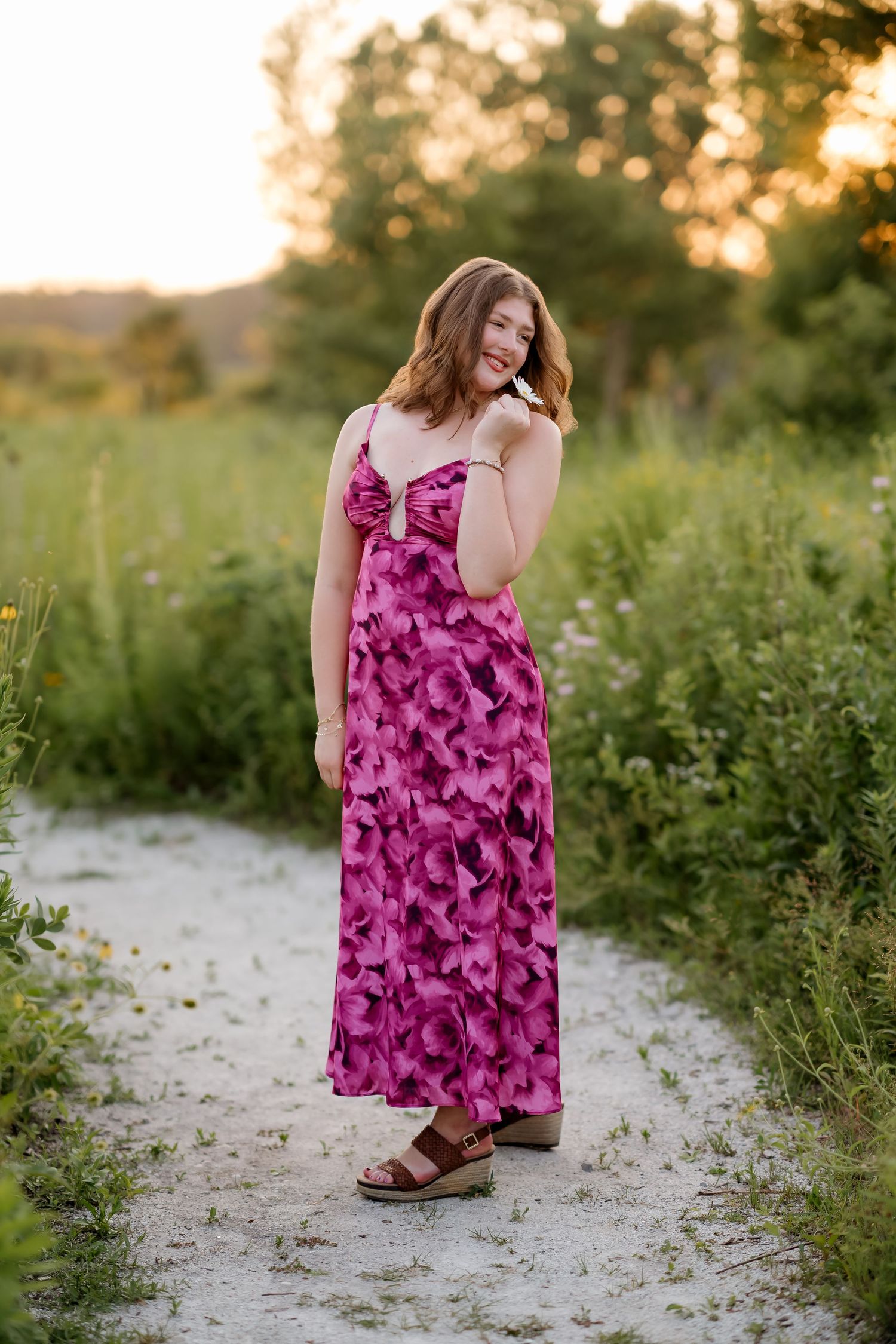 Young woman in pink floral maxi dress and sandals posing on a sandy path surrounded by lush greenery at sunset.
