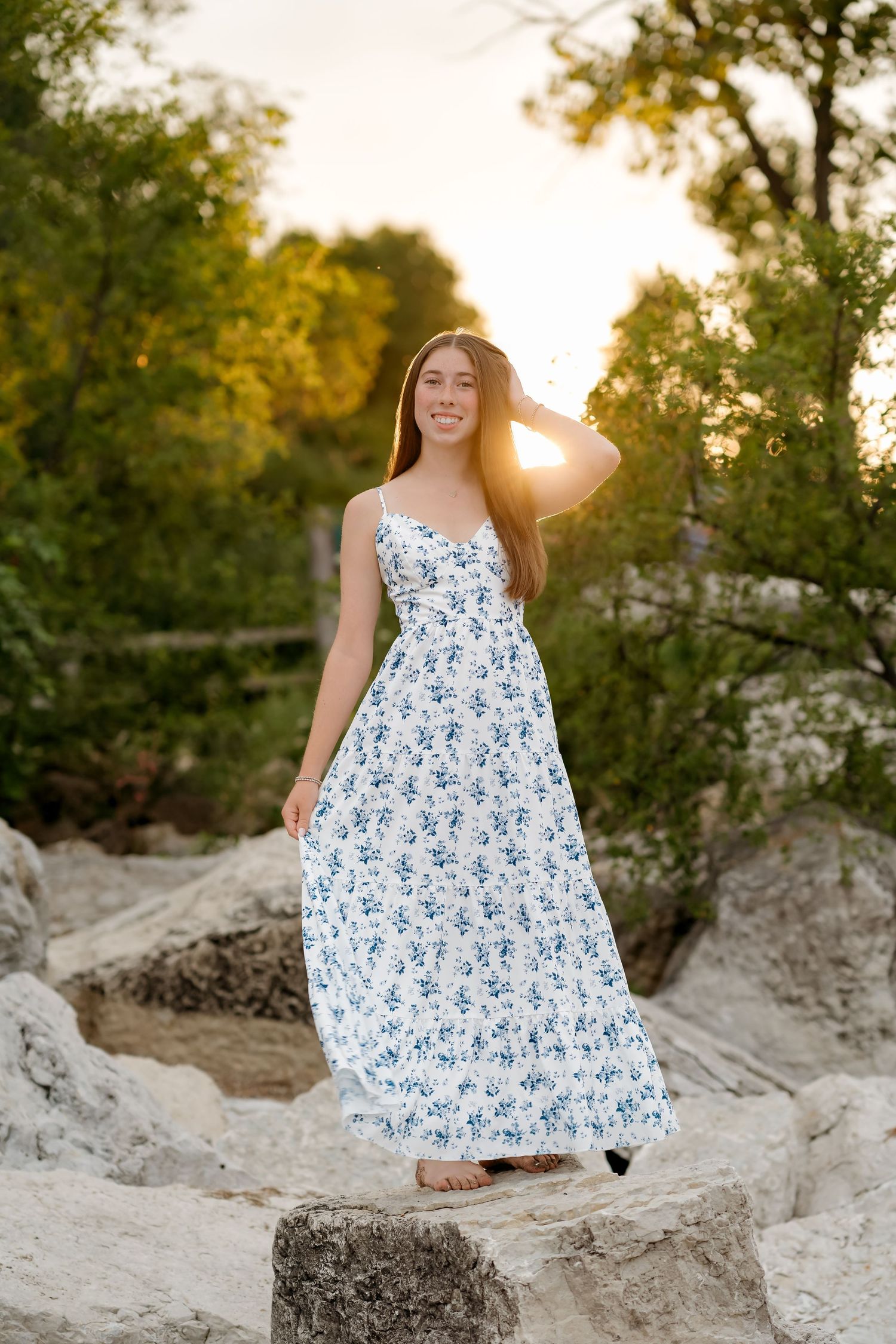 Young woman in white floral maxi dress standing on rocks at sunset with green trees in background.