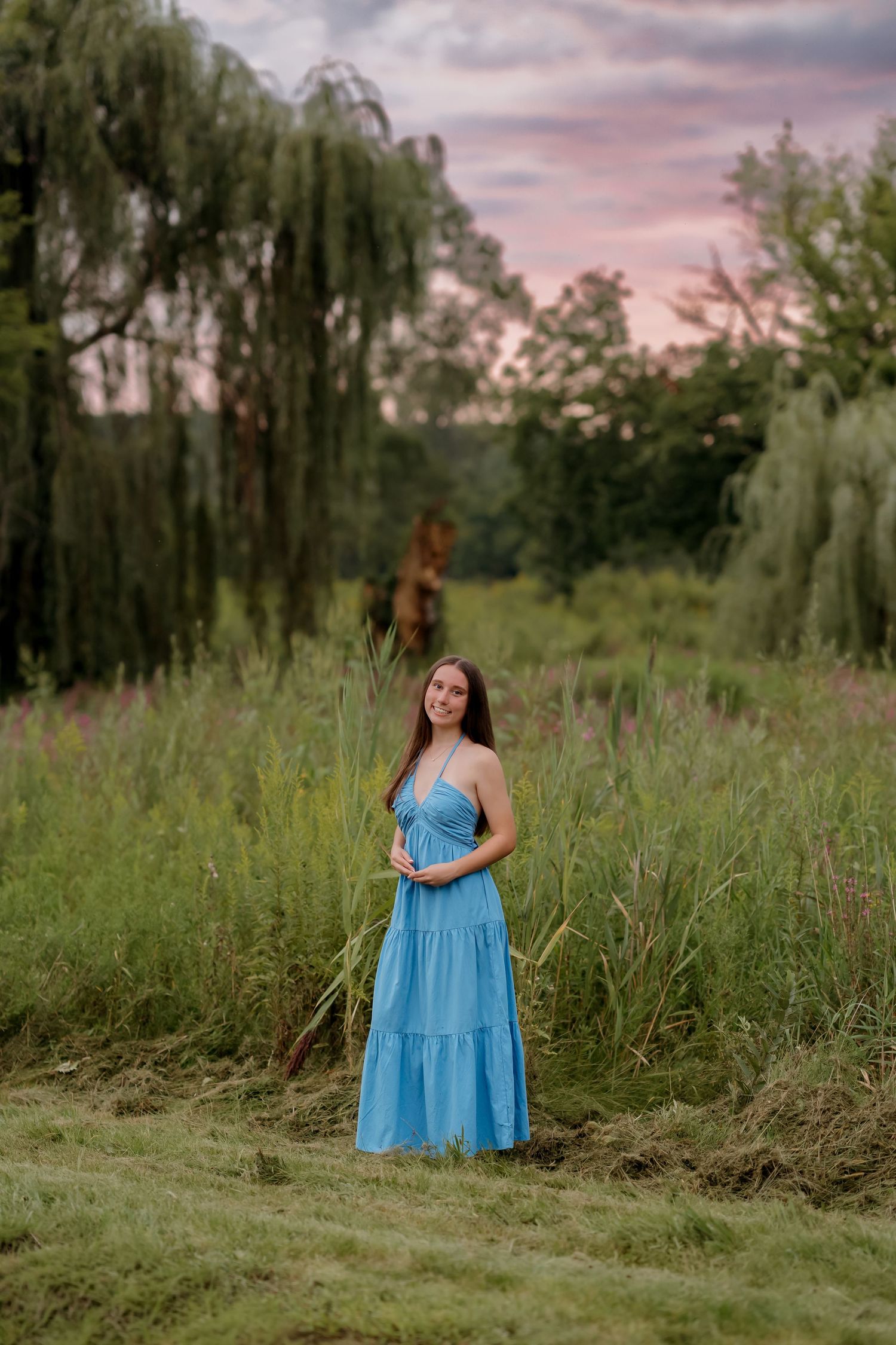 Young woman in blue maxi dress standing in lush green field at sunset with willow trees in background.