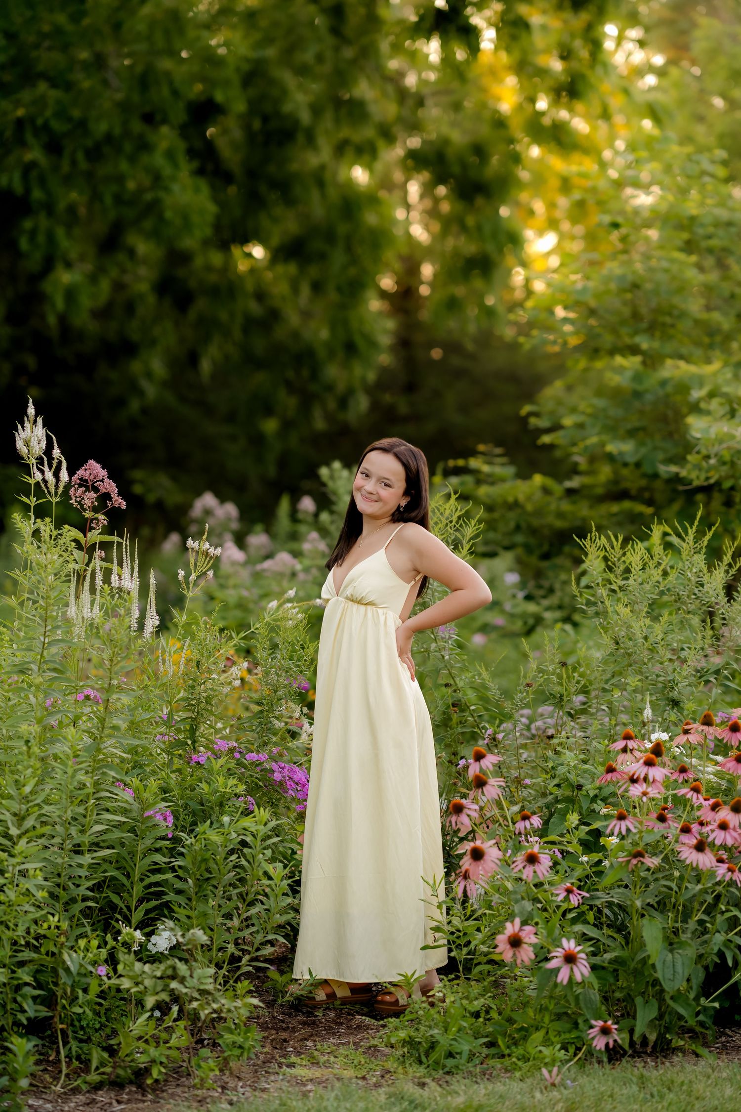 Young woman in cream maxi dress poses among colorful wildflowers in a lush green garden at golden hour.