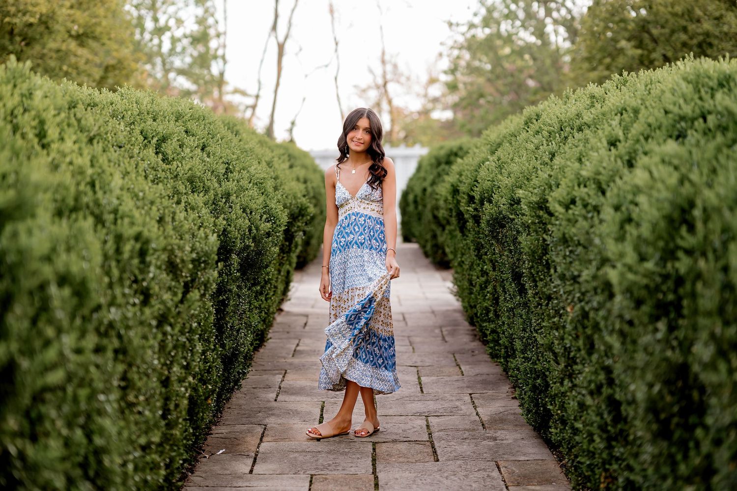 Woman in blue patterned maxi dress walking along a brick path between tall green hedges in a garden.