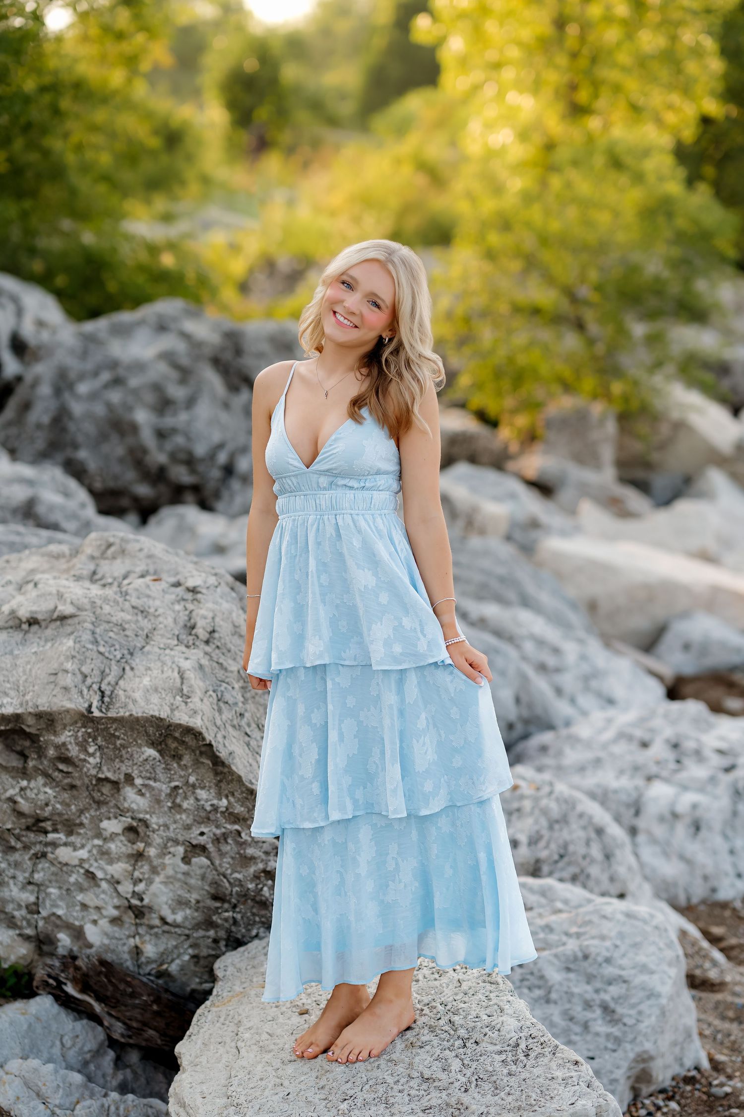 Young blonde woman in a light blue tiered maxi dress standing barefoot on rocks outdoors.