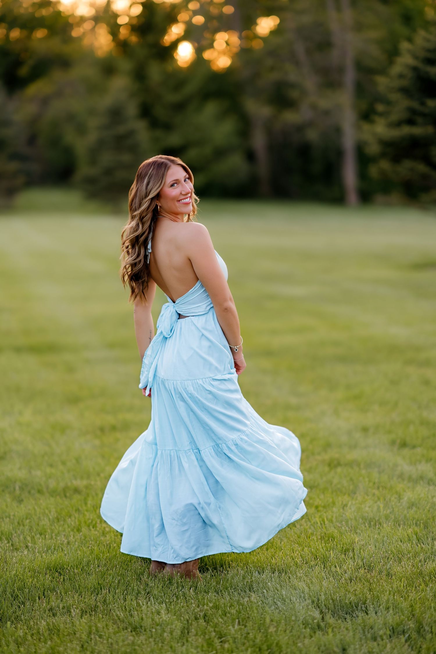 Woman in light blue halter maxi dress twirling on green lawn at golden hour with trees in background.