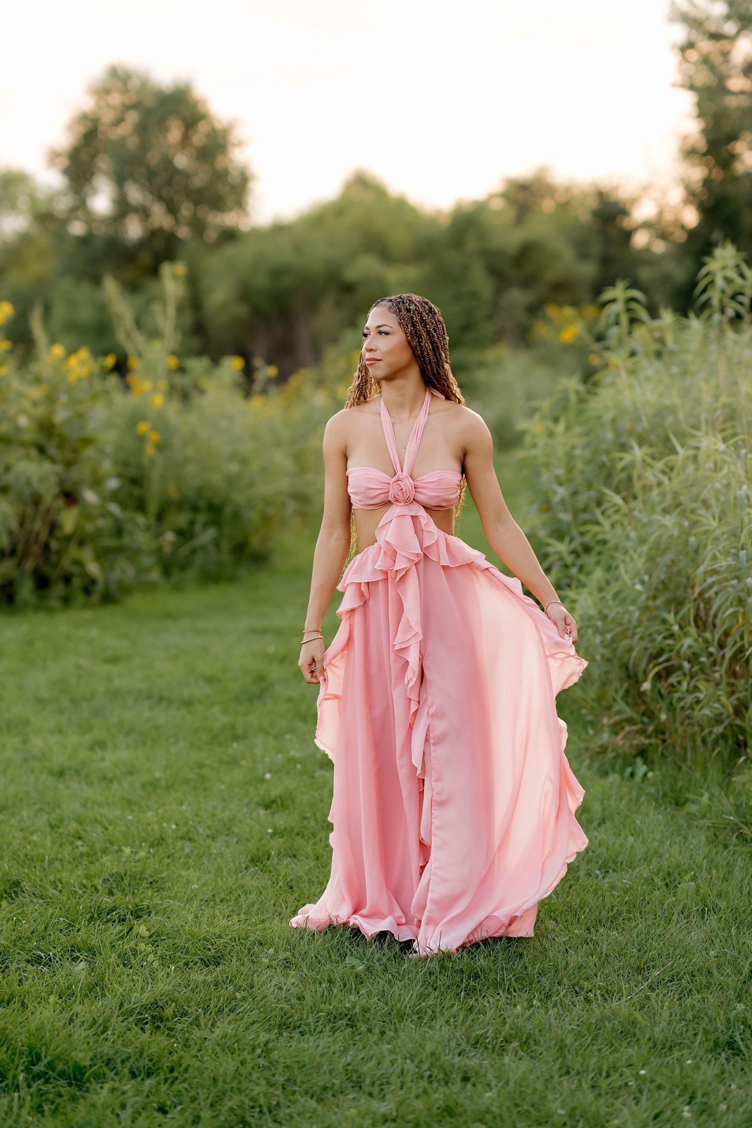 Young woman in flowing pink ruffle dress walking gracefully through lush green garden at golden hour.