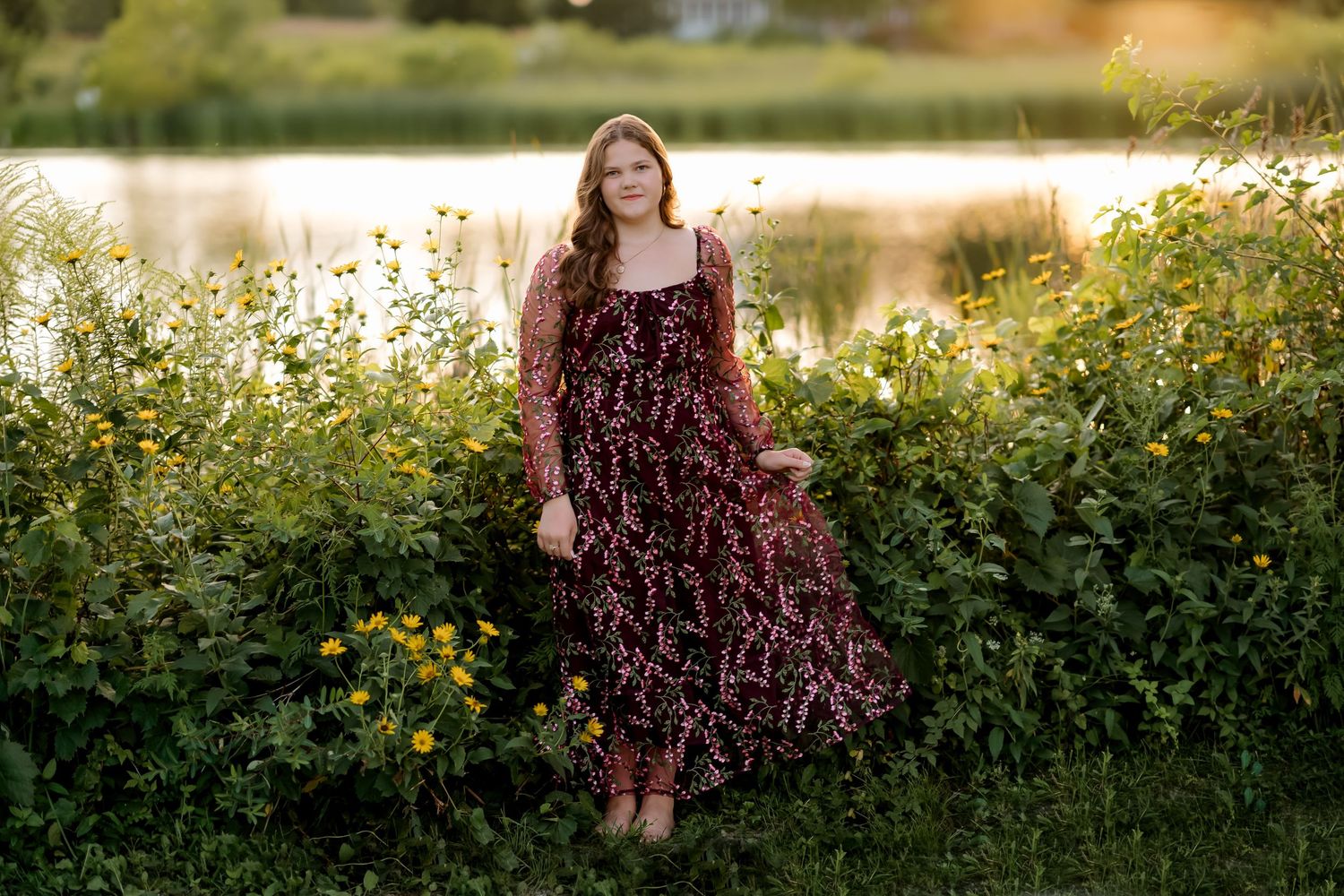 Young woman in floral burgundy maxi dress standing among wildflowers near a sunlit pond at golden hour.