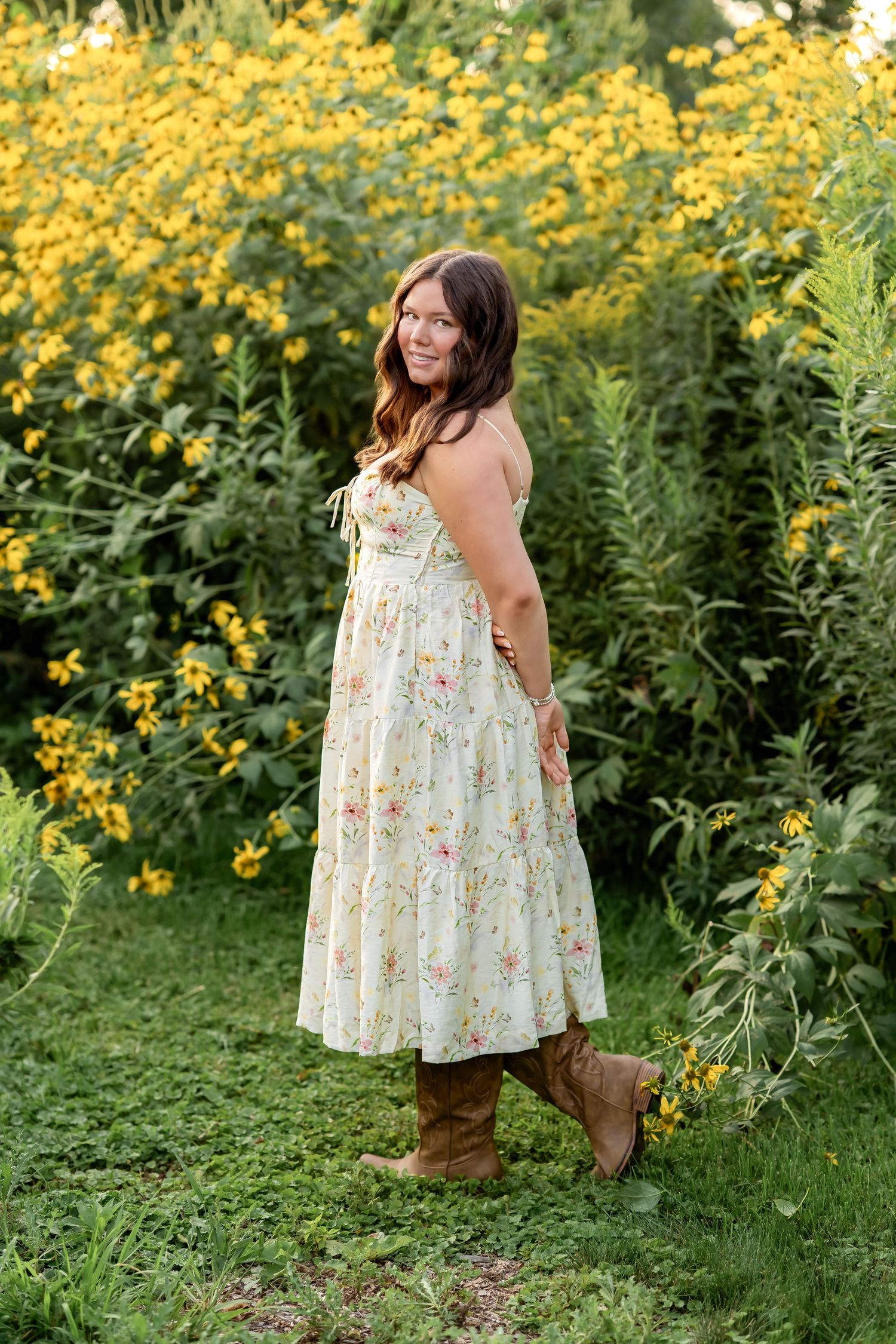 Young woman in floral midi dress and brown boots standing among yellow wildflowers in a lush garden.