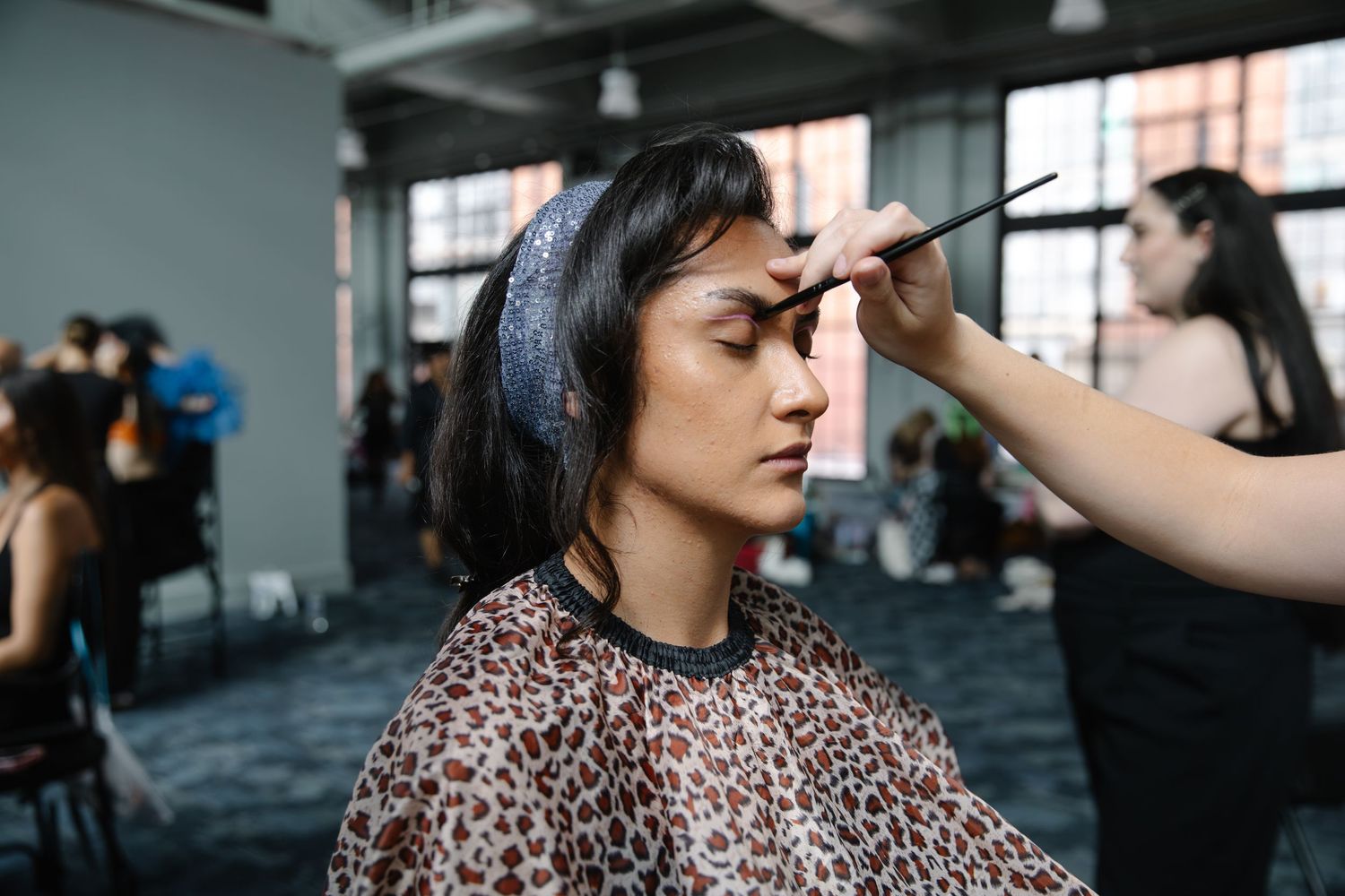 A makeup artist applies eyebrow makeup to a model wearing a leopard print cape at a fashion show backstage.