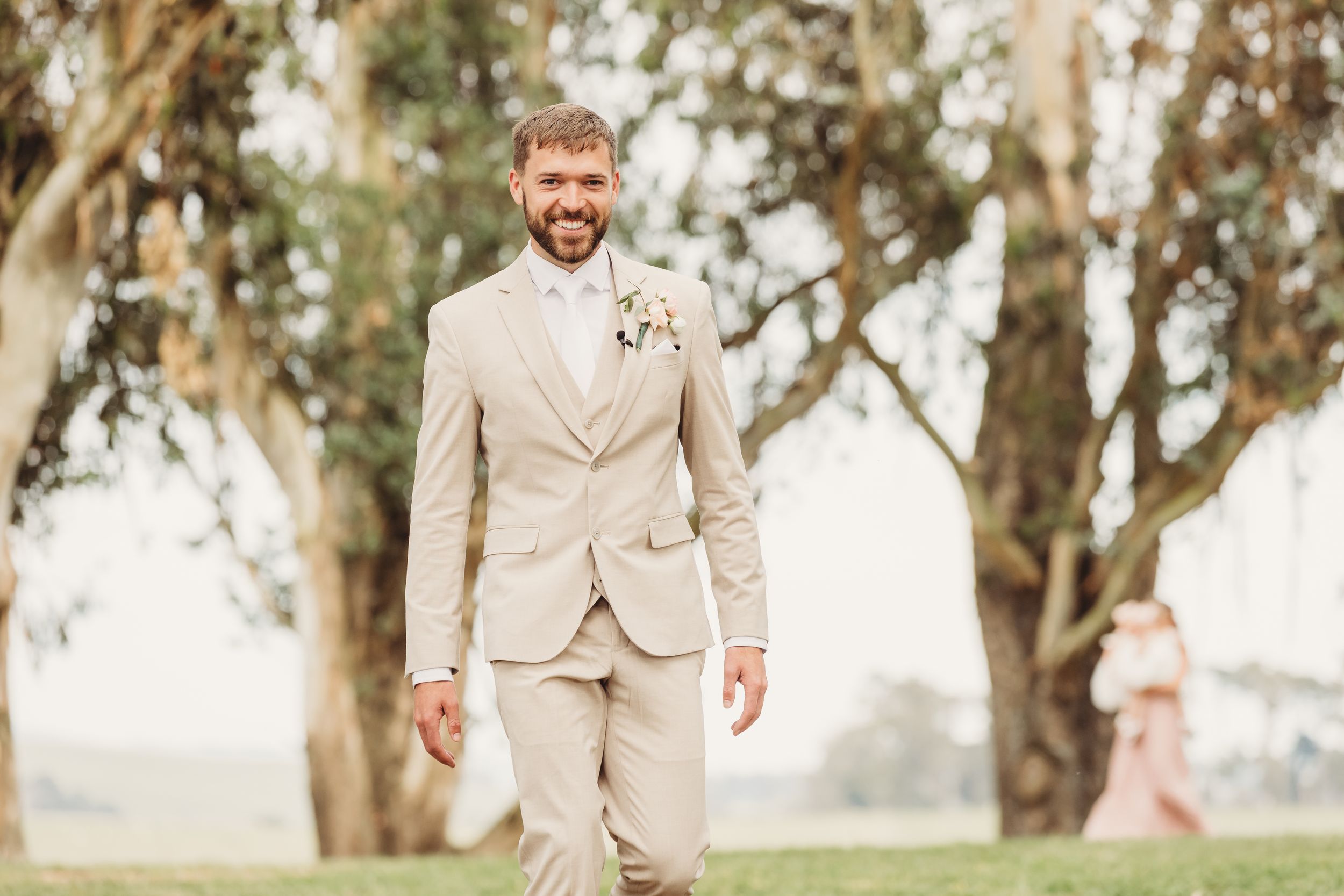 Smiling groom in beige suit with boutonniere walks outdoors at wedding venue surrounded by trees.