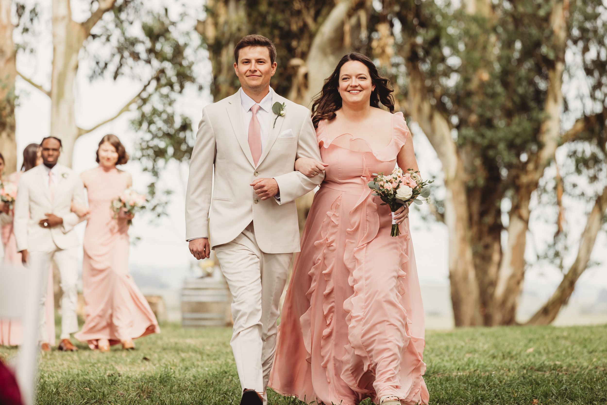 Bride in pink ruffled gown walks with groomsman in tan suit at outdoor wedding ceremony.
