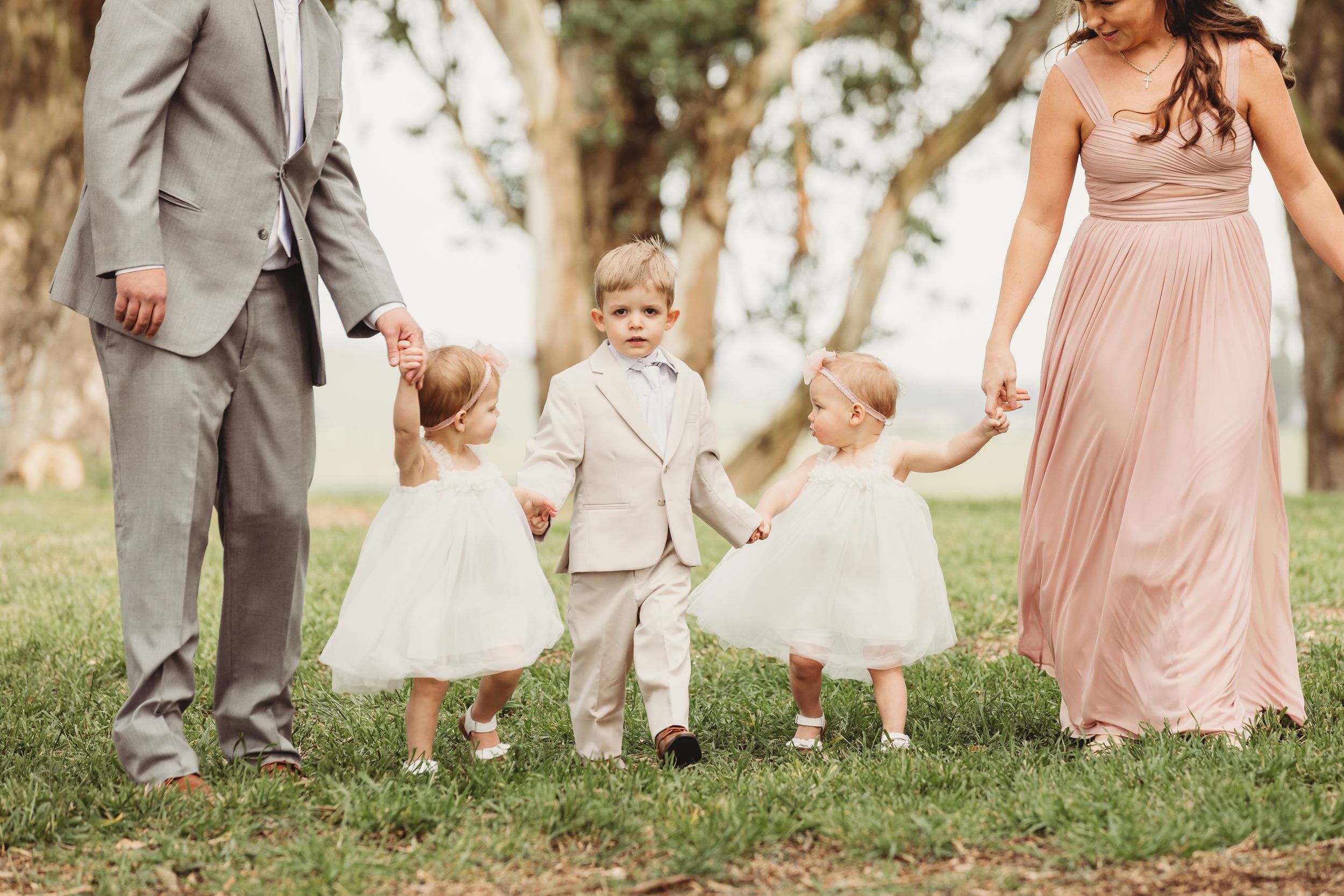 Family in formal attire with two flower girls in white dresses and a ring bearer walking outdoors on grass.