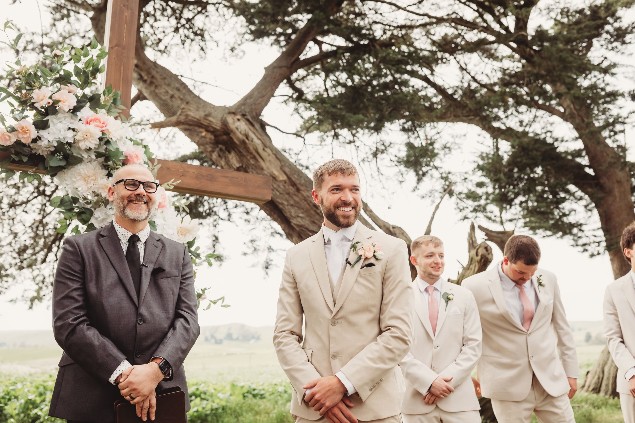 Groom smiling at altar during outdoor wedding ceremony with groomsmen in tan suits and pink ties.