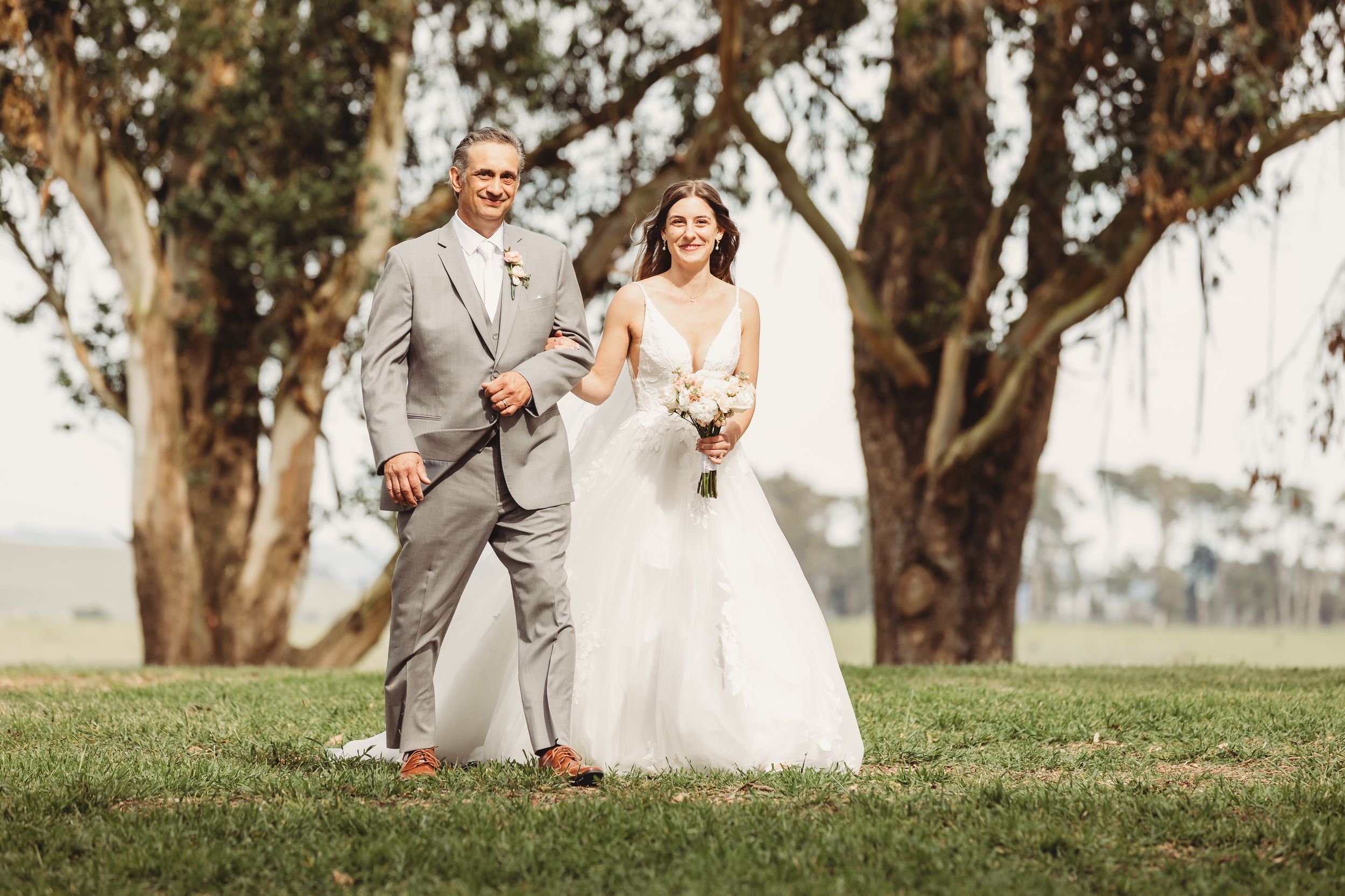 Bride in white gown and groom in gray suit walk together outdoors near large trees on wedding day.
