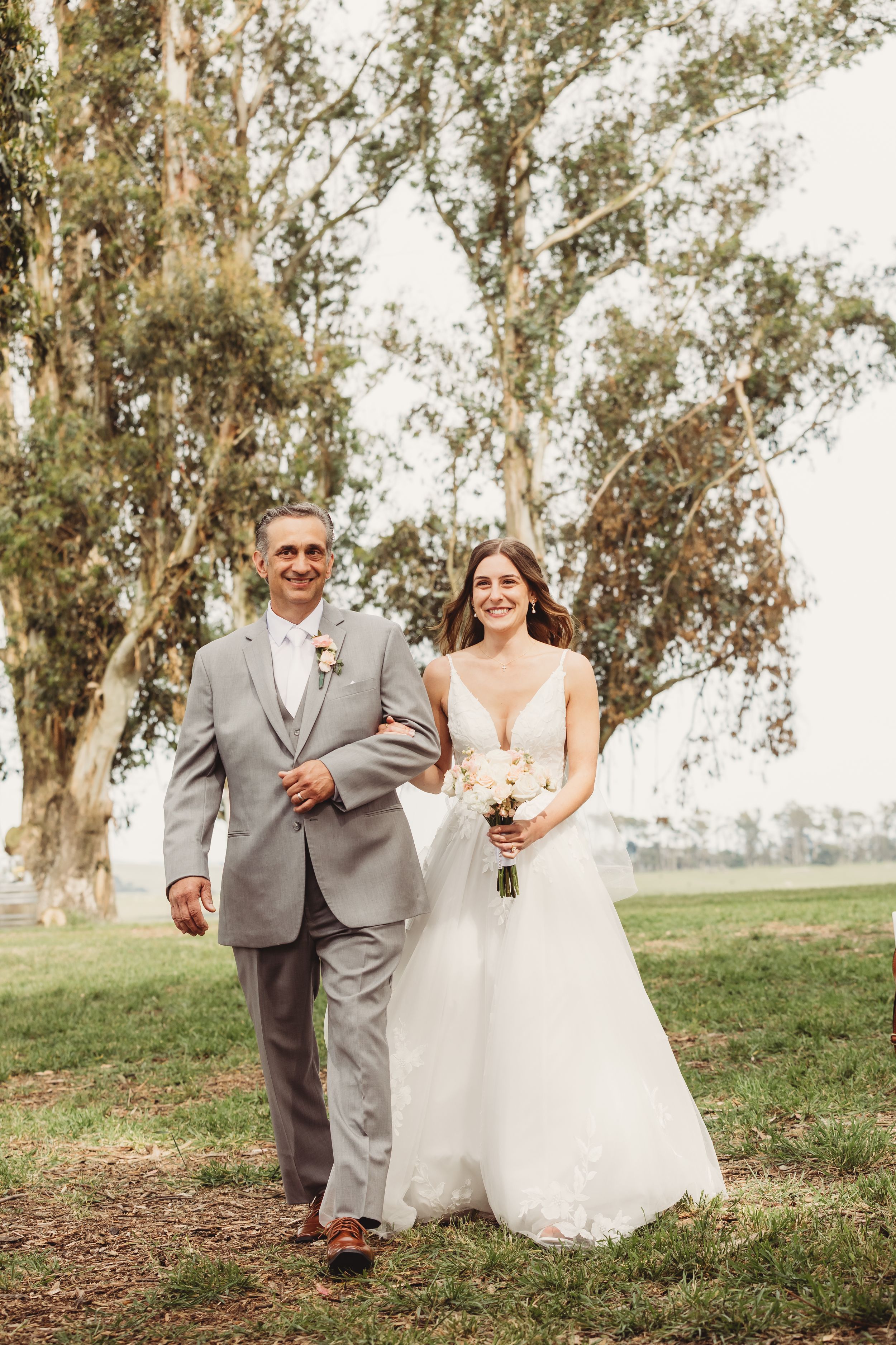 Bride in white gown holding bouquet walks arm-in-arm with man in gray suit outdoors near tall trees.