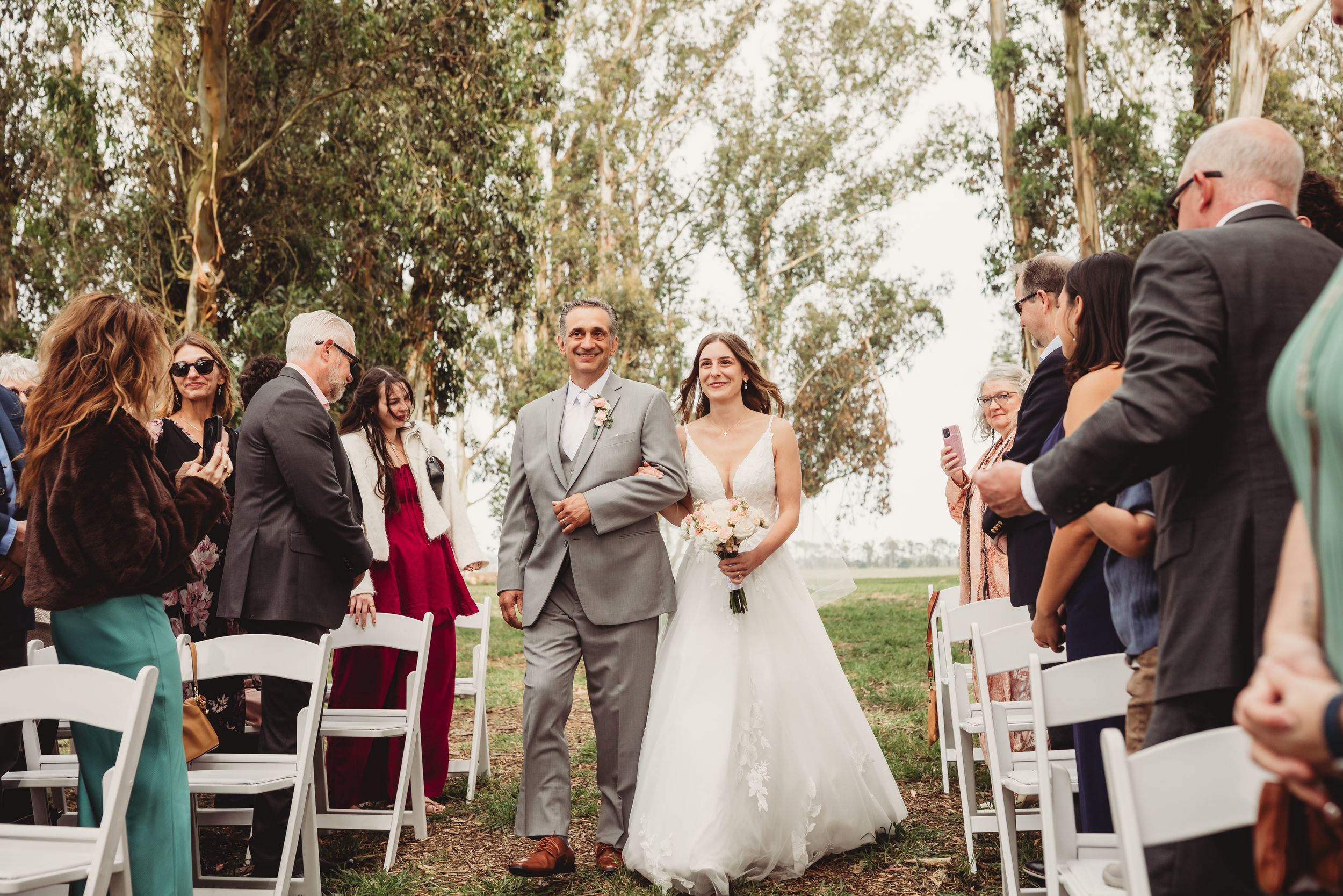 Bride walks down outdoor aisle with escort, guests cheering, white chairs, trees in background at wedding ceremony.