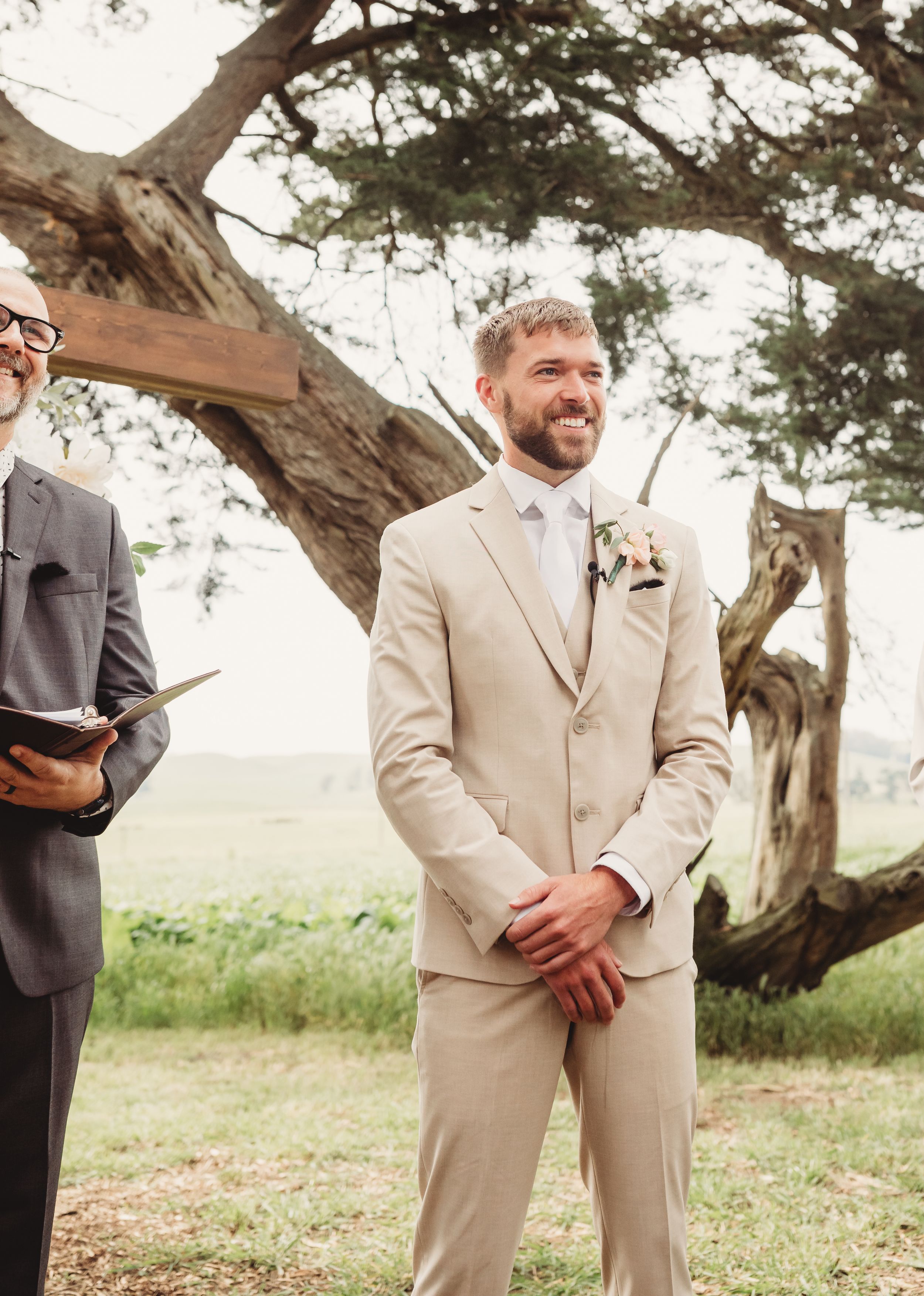 Smiling groom in beige suit with floral boutonniere standing at outdoor wedding ceremony beside officiant.