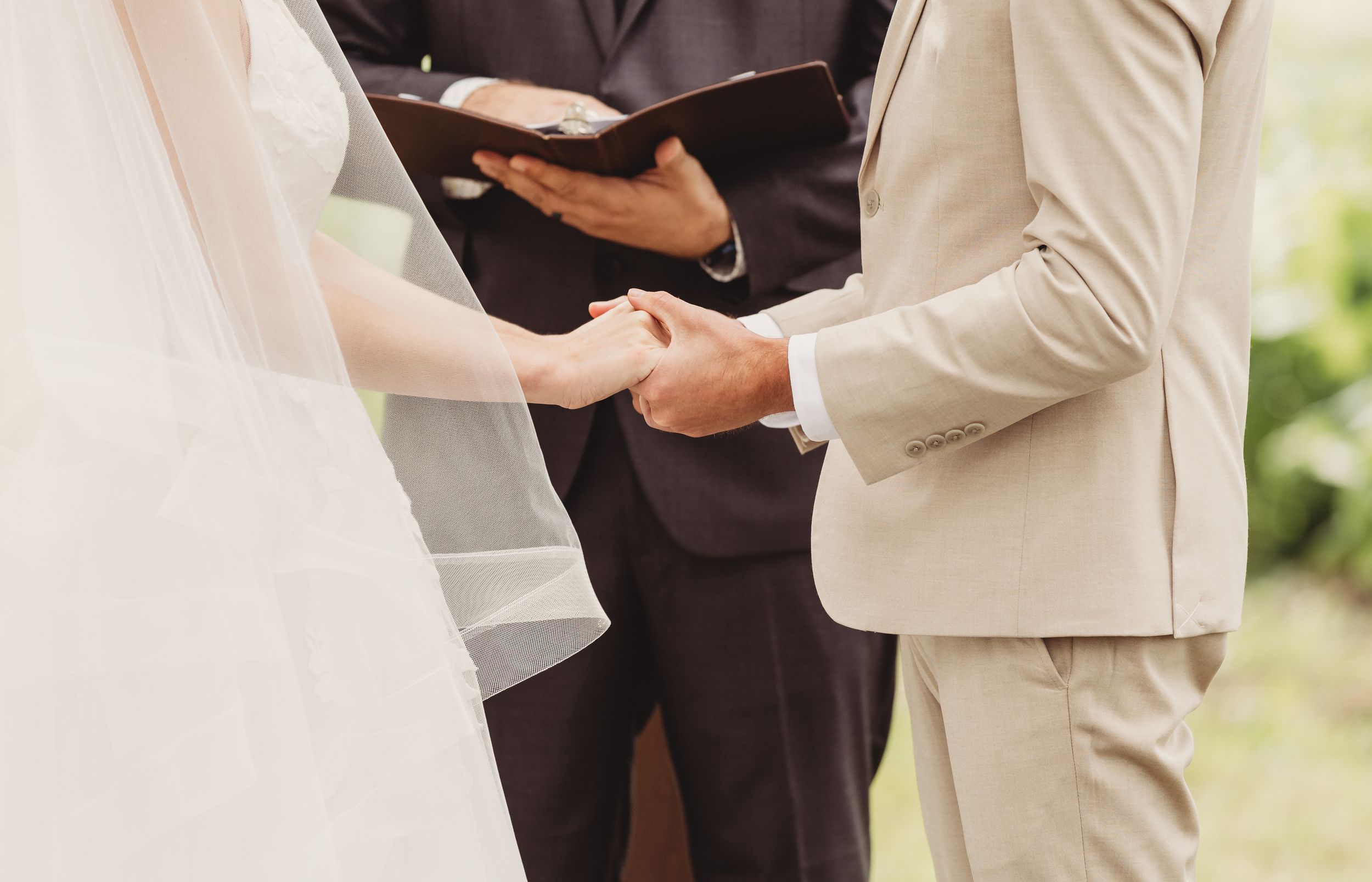Bride and groom holding hands during wedding ceremony while officiant reads from book outdoors.