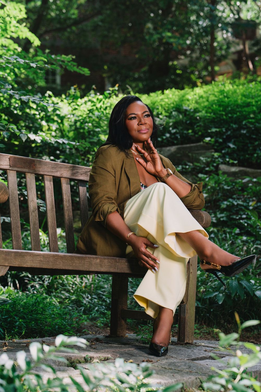 Woman in olive blazer and cream skirt sits gracefully on wooden bench surrounded by lush green garden foliage.