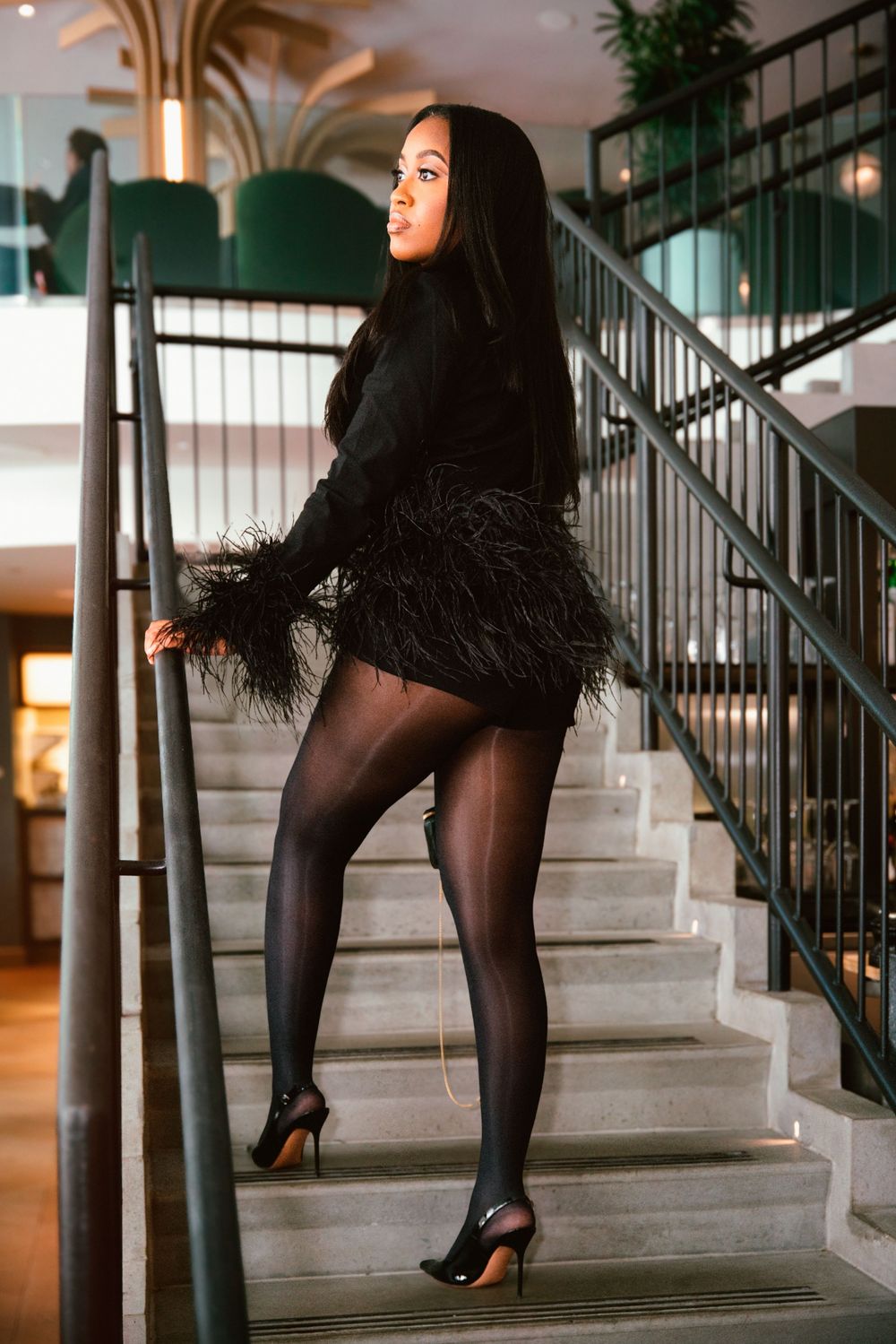 Woman in black feather mini dress, sheer tights, and heels posing elegantly on marble staircase.