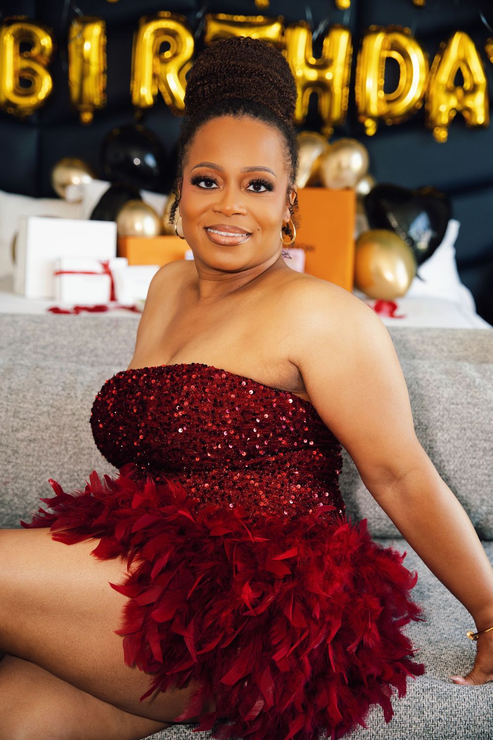 Woman in red sequin feather dress smiling at birthday party with gold balloon letters and black and gold decor.