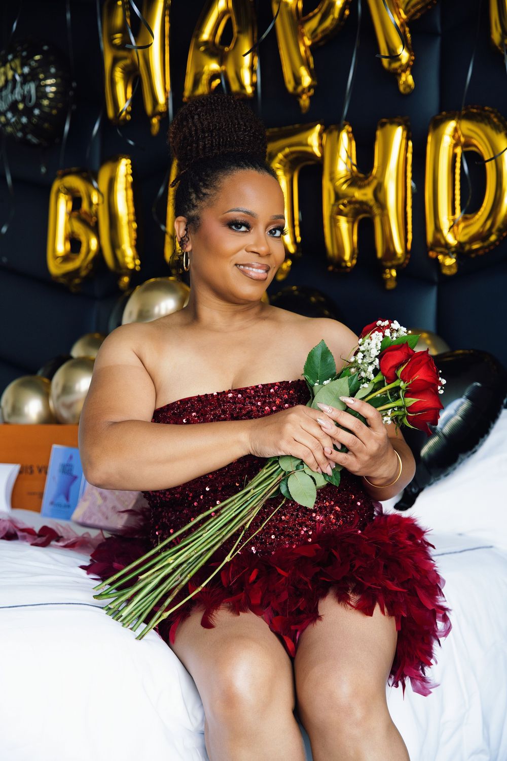 Woman in red sequin dress holding roses, celebrating birthday with gold balloon decorations.